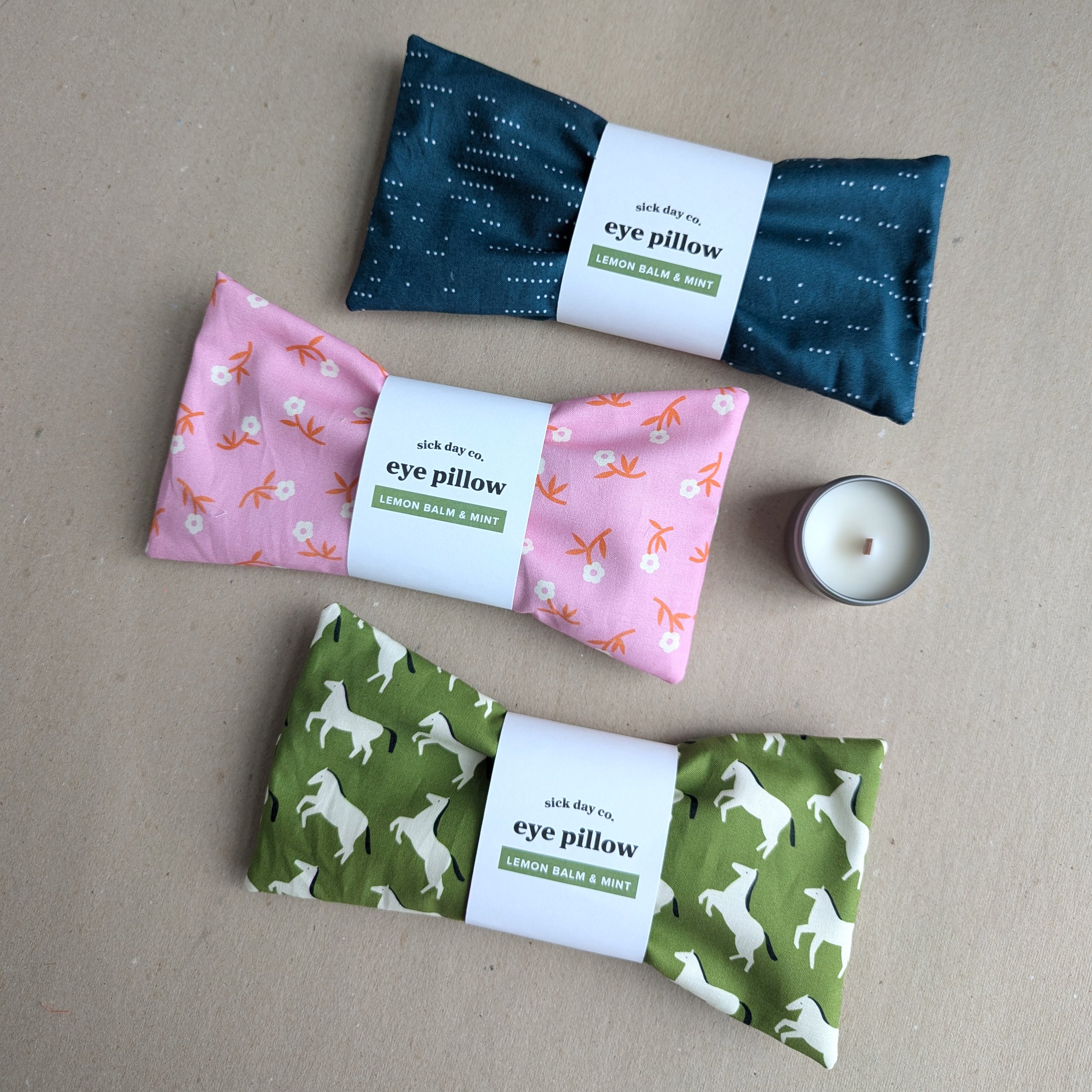 Three folded eye pillows with colorful textiles and white labels on a beige surface, labeled 'sick day co.' and 'eye pillow' with scents 'Lemon Balm & Mint'.