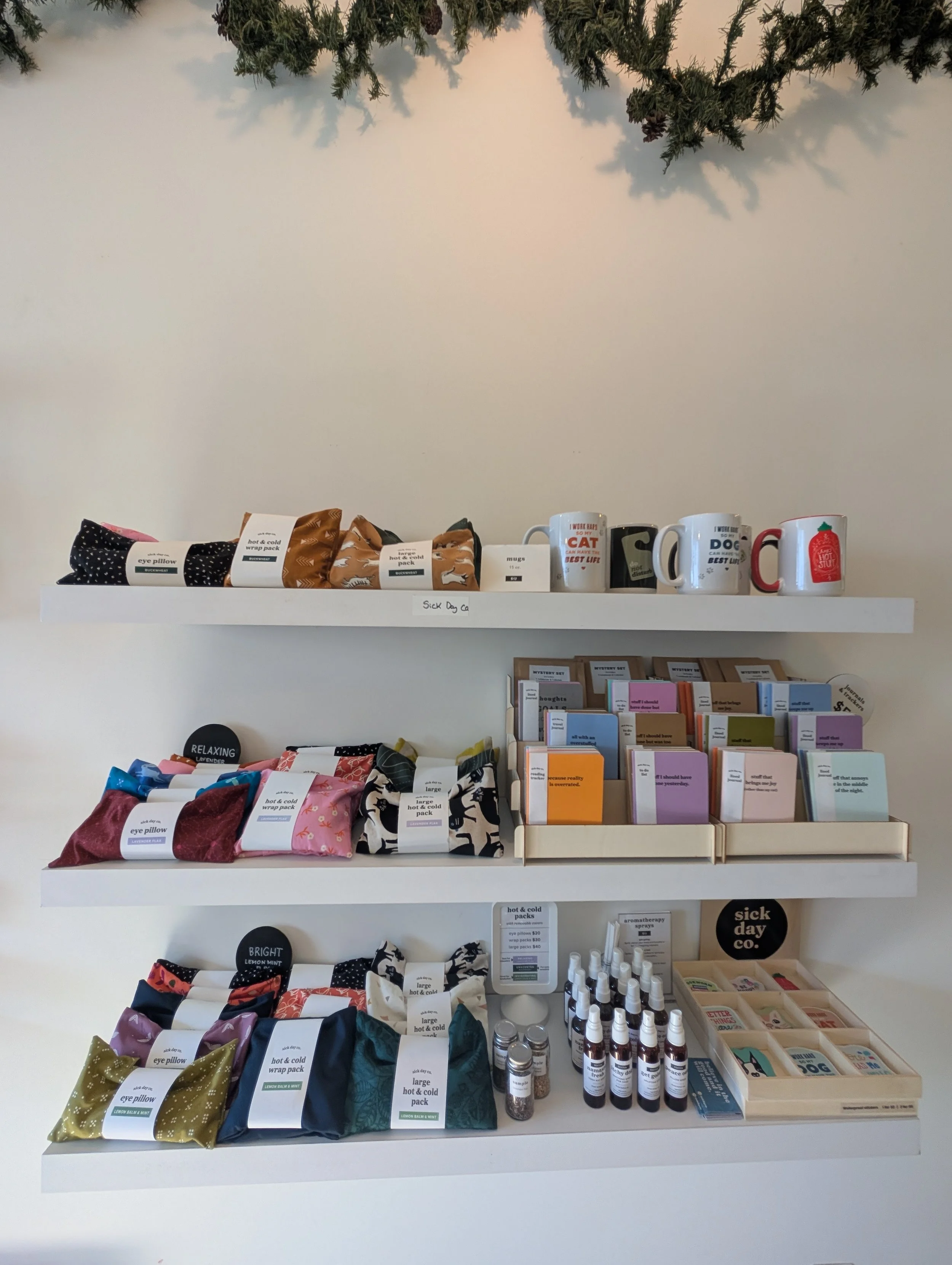 Display of various products on shelves, including eye pillows with polka dot and floral patterns, hot and cold wrap packs with lemon and mint designs, character-themed mugs, and small bottles, with some books or pamphlets in a wooden tray below. A small sign reads 'sick day co.'.