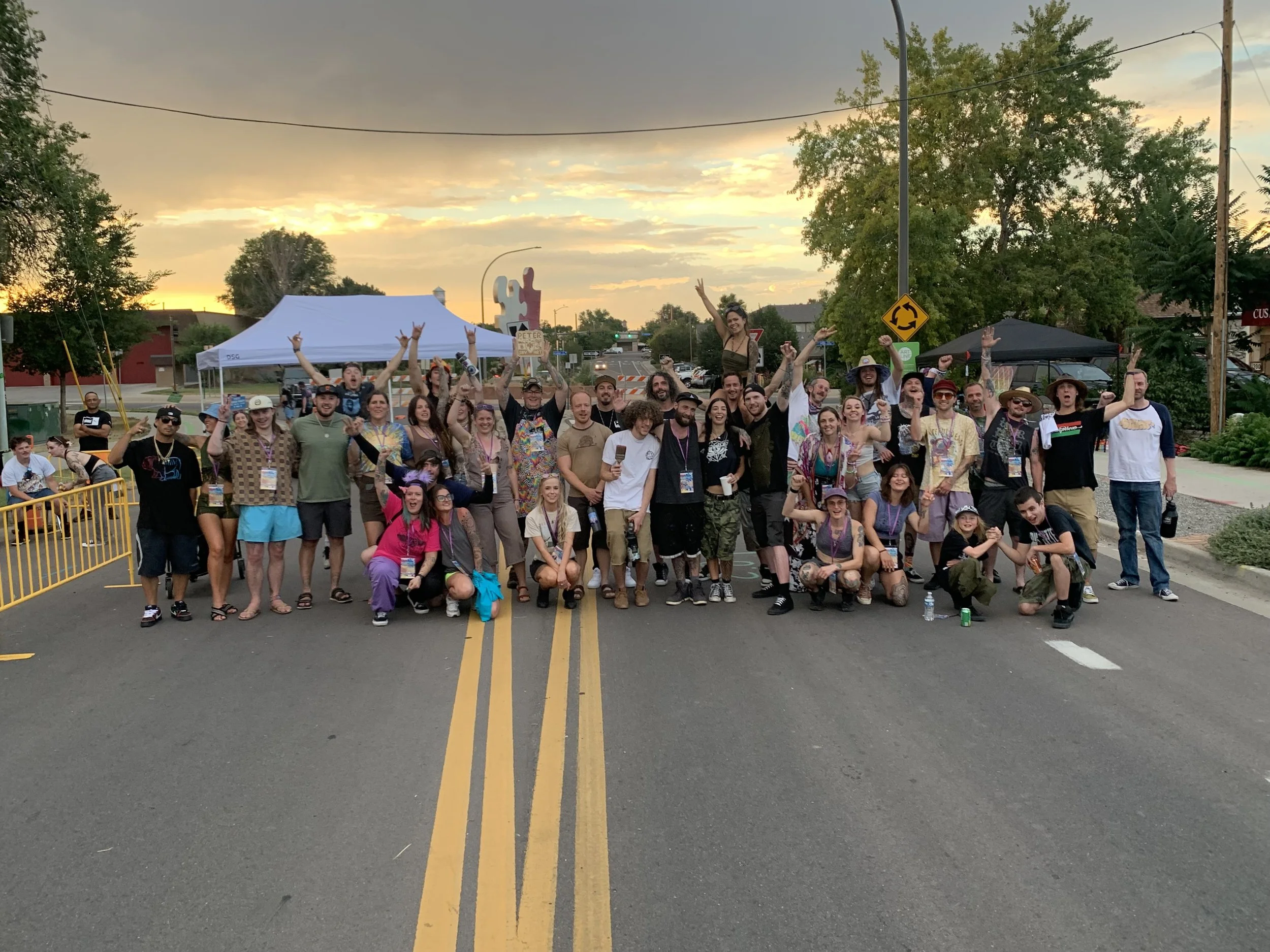 A large group of people posing for a photo on a city street at sunset. The group includes men, women, and children, some raising their hands and making gestures. There is a white canopy tent and a yellow roundabout sign in the background, as well as trees and buildings.