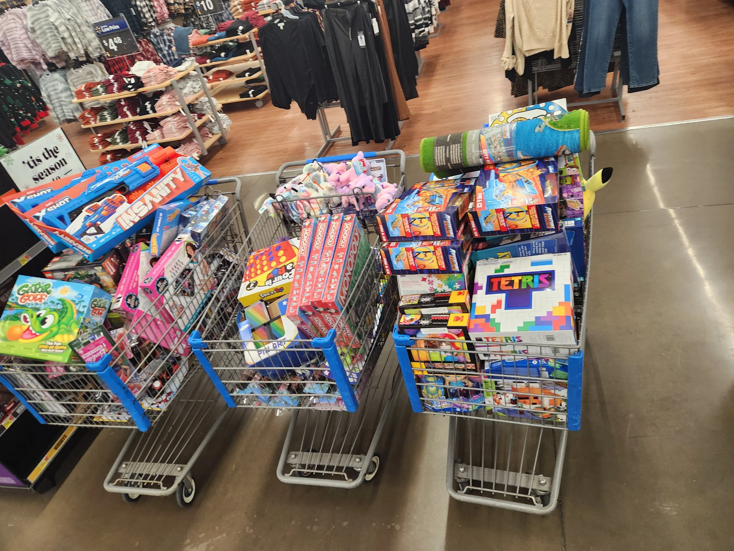 Three shopping carts filled with toys, including board games, plush toys, and puzzles, in a store aisle with clothing displays in the background.