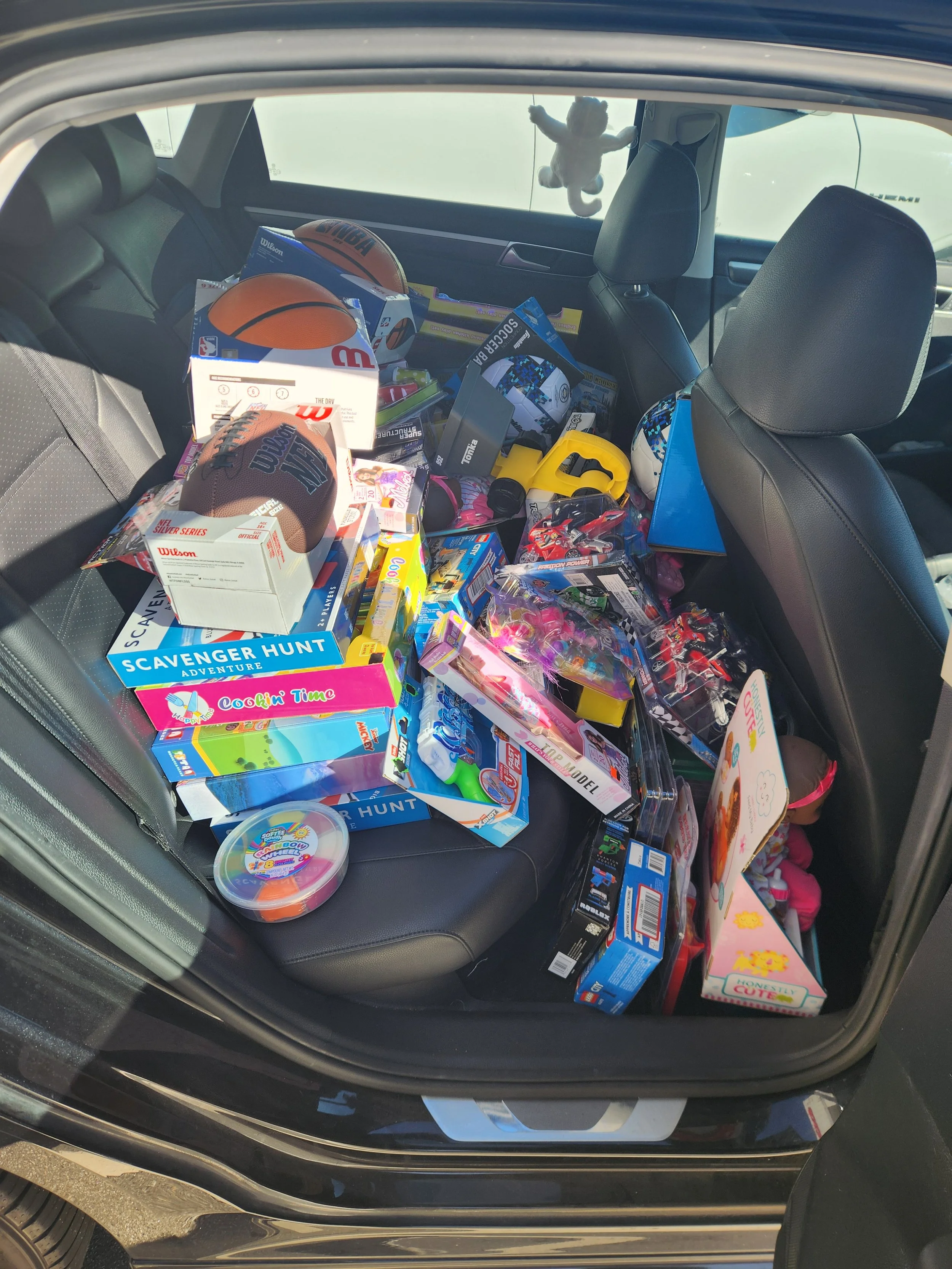 Car trunk filled with toys, games, and plush dolls, including sports balls, board games, and children's toys.
