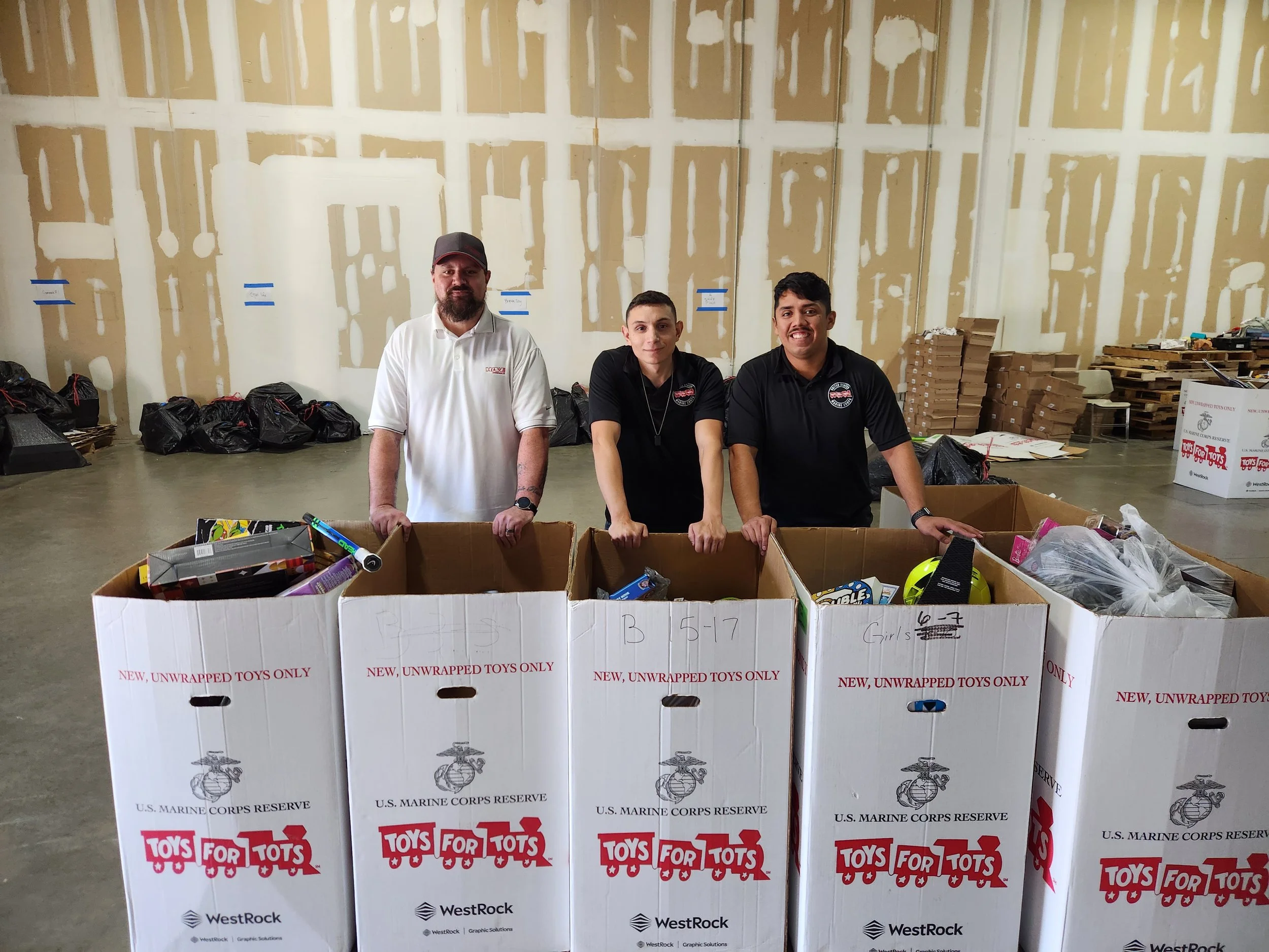 Three men standing behind large boxes labeled 'Toys for Tots' in a warehouse with a partially finished drywall wall in the background.