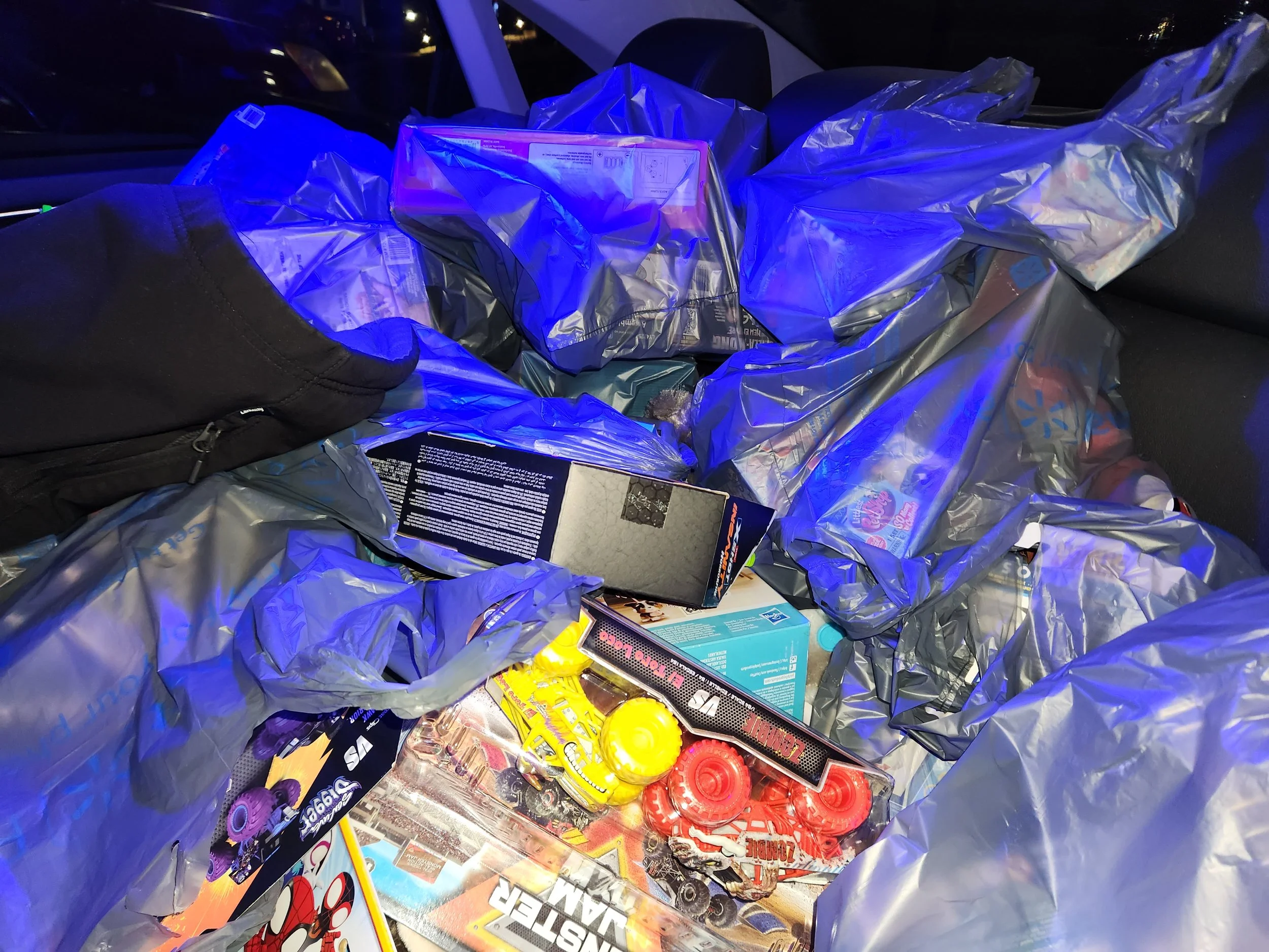 A pile of plastic shopping bags containing various items, including board games, toys, and candy, inside a vehicle at night.