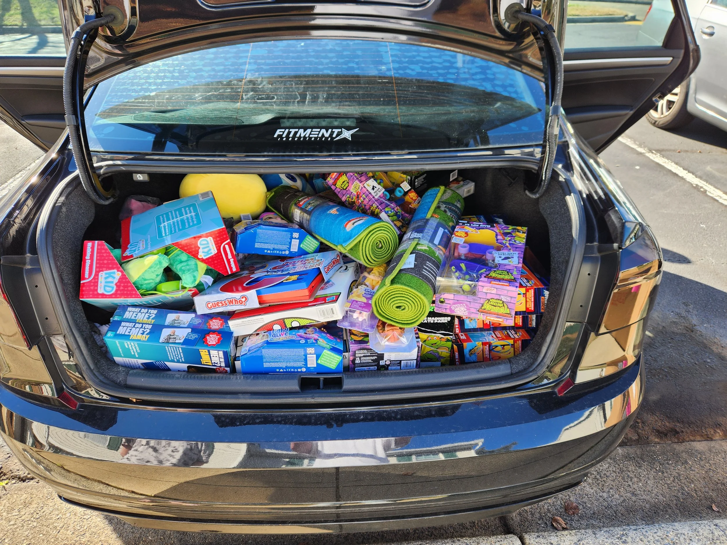 The trunk of a black car is filled with various toys, including board games, a foam mat, a basketball, and other miscellaneous items, with the car parked in an outdoor parking lot.