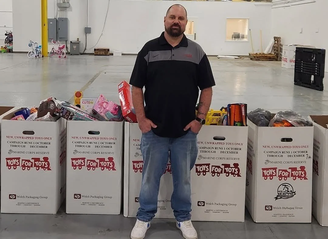 A man standing in a warehouse with boxes of toys for a Toys for Tots charity drive.