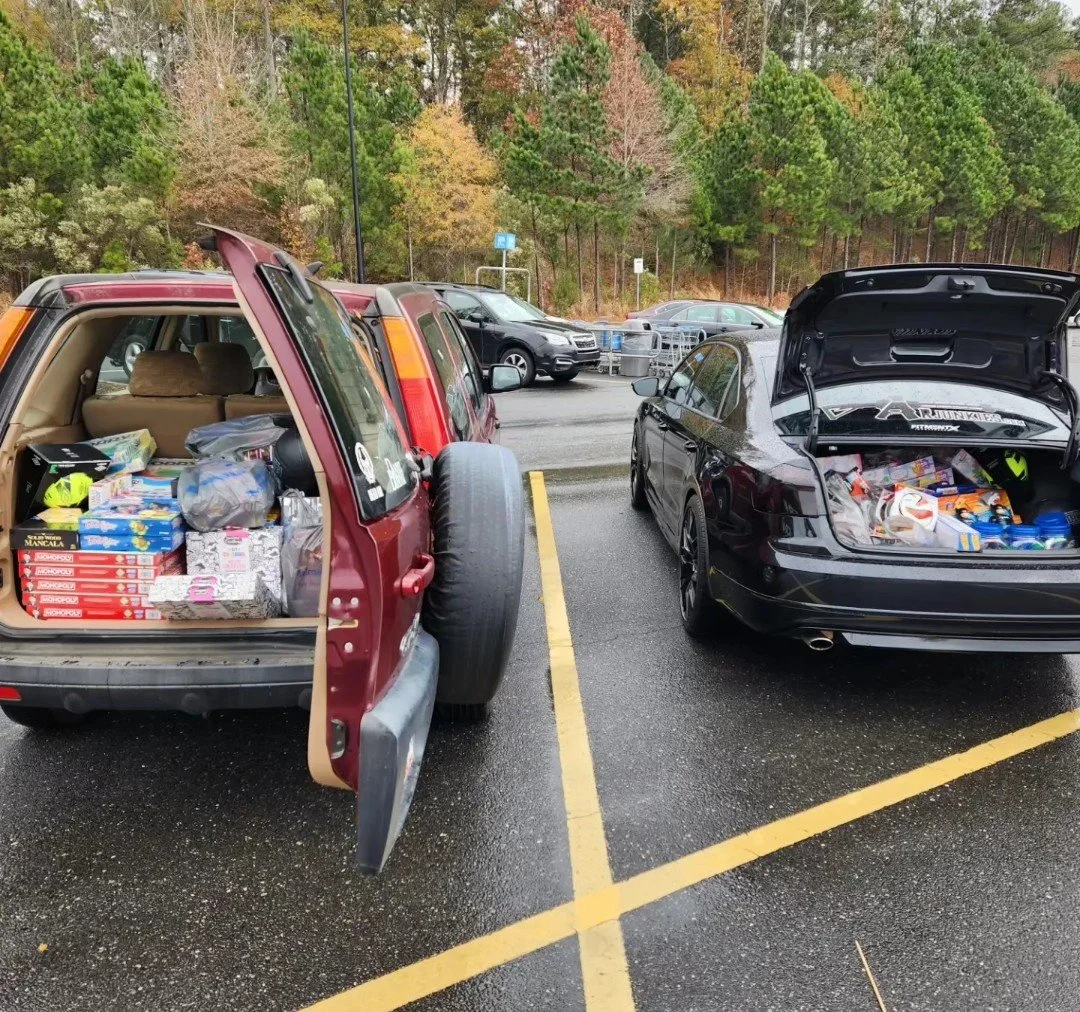 Two cars in a parking lot with their trunks open, filled with groceries. The left car is a maroon SUV with a spare tire mounted on the back. The right car is a black sedan. In the background, there are trees and other parked cars.