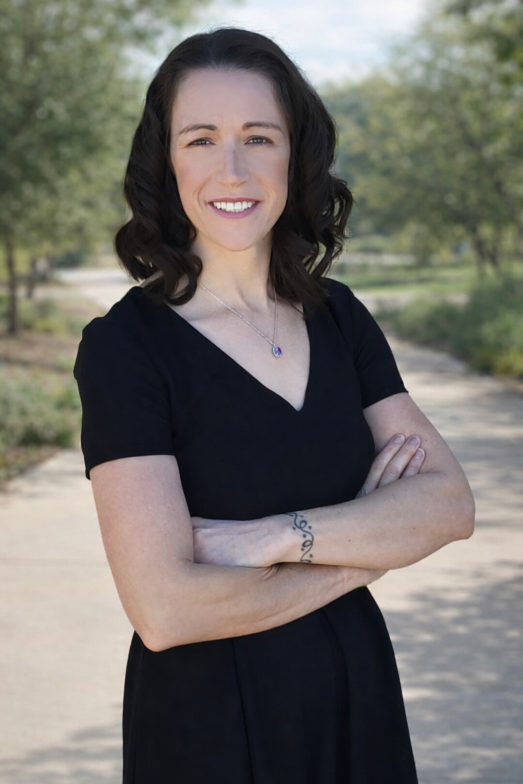 A woman with dark brown wavy hair wearing a black V-neck dress, standing outdoors with trees in the background, smiling with her arms crossed.