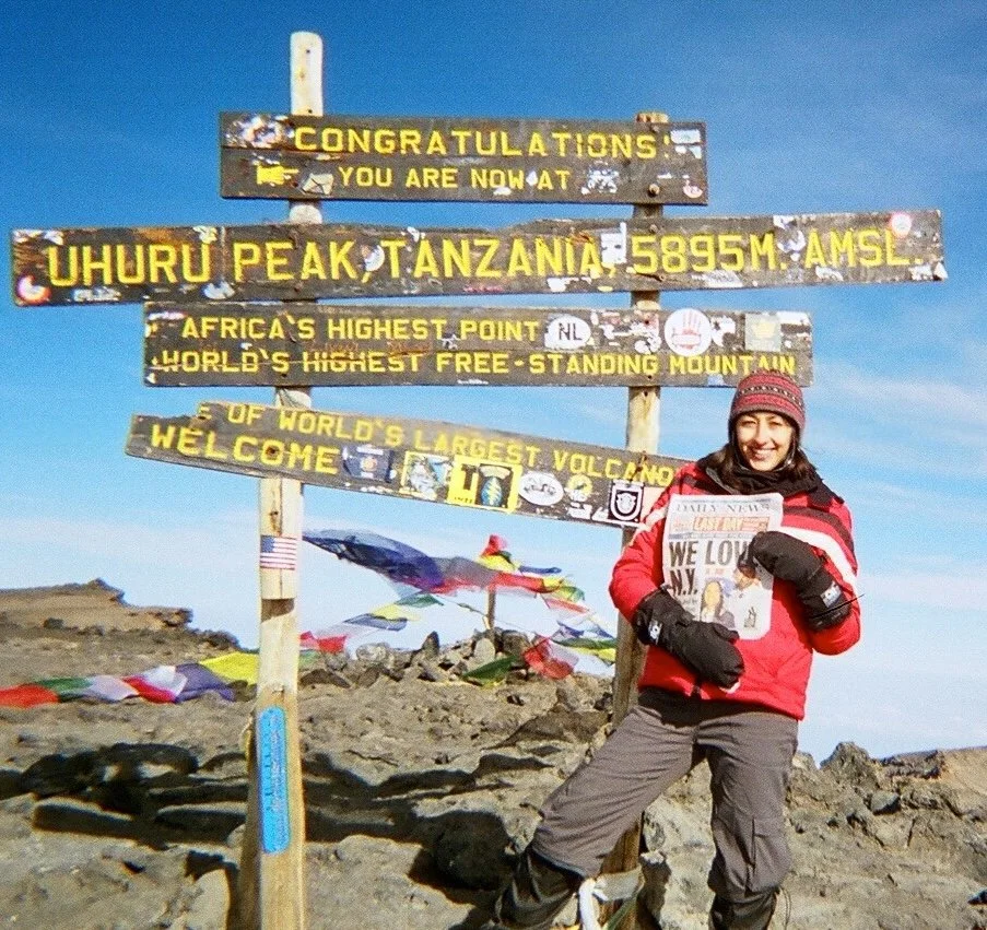 A woman on Top of Uhuru Peak in Tanzania holding a newspaper, standing beside a sign marking the summit of Mount Kilimanjaro at 5895 meters, with clear sky and some prayer flags in the background.