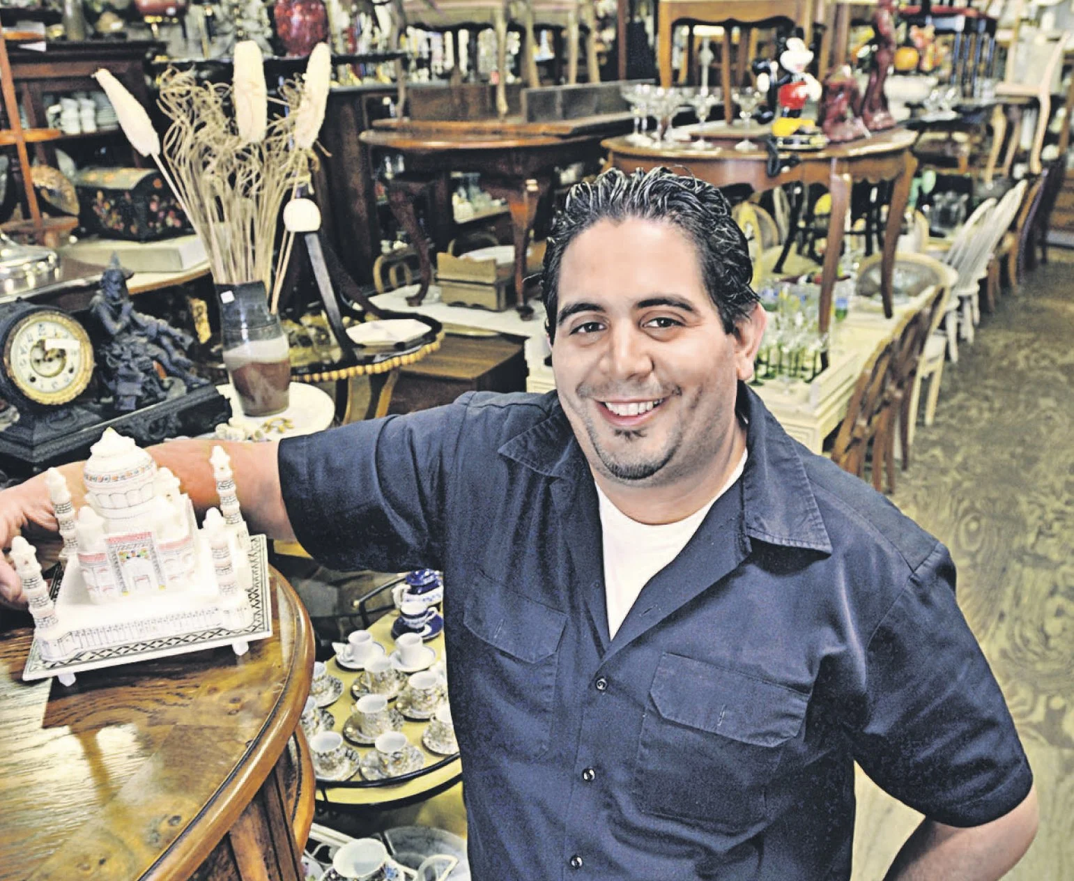 A smiling man with dark, slicked-back hair wearing a navy blue shirt, sitting at a table with various decorative items and collectibles, surrounded by furniture and ceramics in an antique shop or flea market setting.