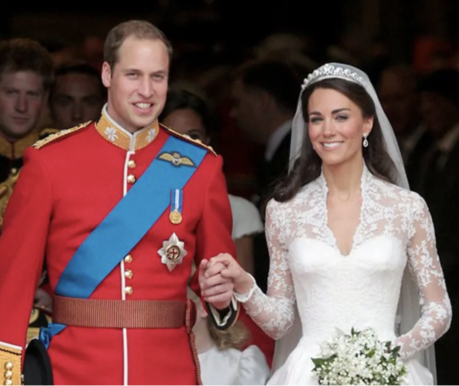 A bride and groom at a wedding, holding hands. The groom is wearing a red military uniform with medals and a blue sash, and the bride is in a lace wedding dress with a veil, holding a bouquet of white flowers.