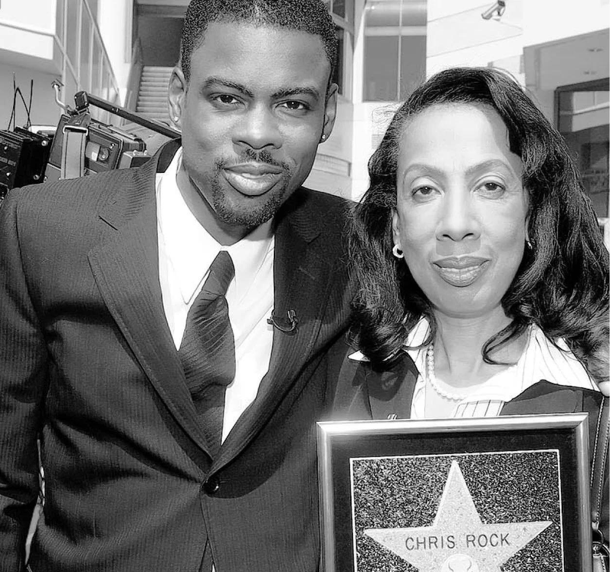A man and woman posing together, the woman holding a plaque with the name 'Chris Rock' on it, in an indoor setting with a staircase and cameras visible in the background.