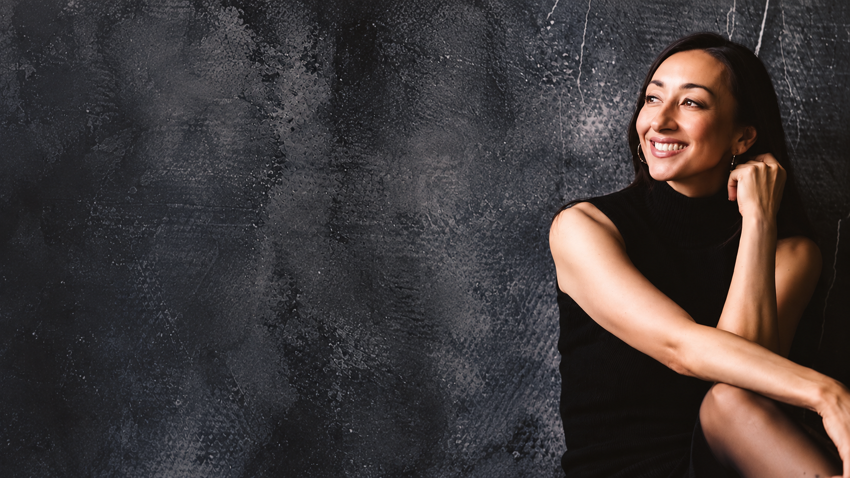 Eloise Parker wearing black sleeveless dress with long dark hair smiling, sitting against a textured dark wall.