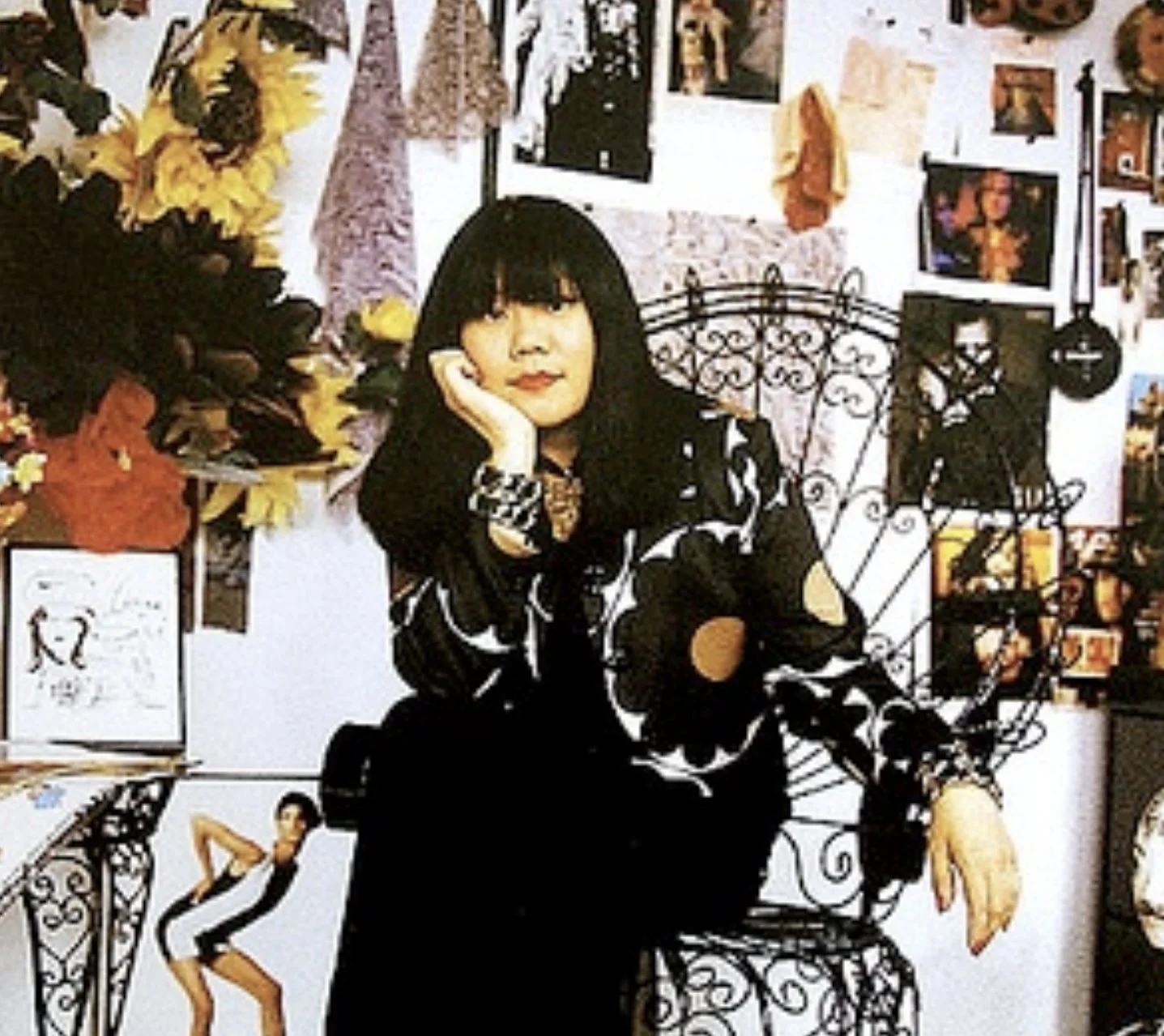 A woman with black hair and bangs, sitting on a decorative black metal chair, resting her chin on her hand, and wearing a black and white patterned outfit, in a room decorated with photographs, artwork, and colorful paper flowers on the wall.