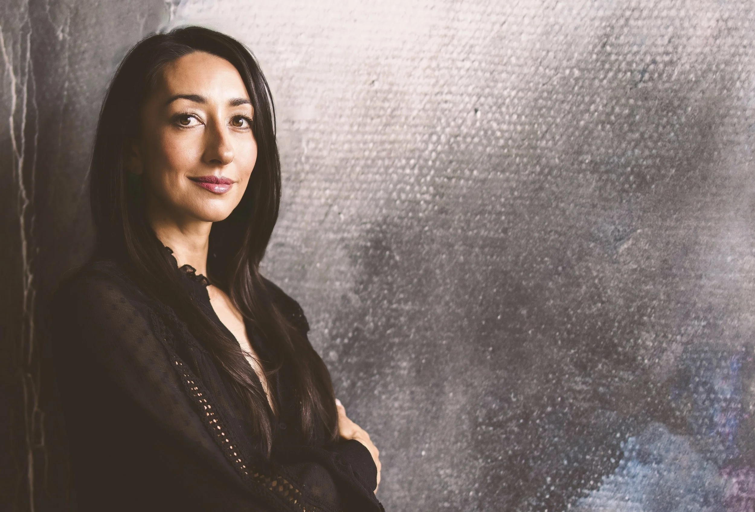 Eloise Parker with long dark hair, wearing a black blouse, standing beside a textured gray wall.