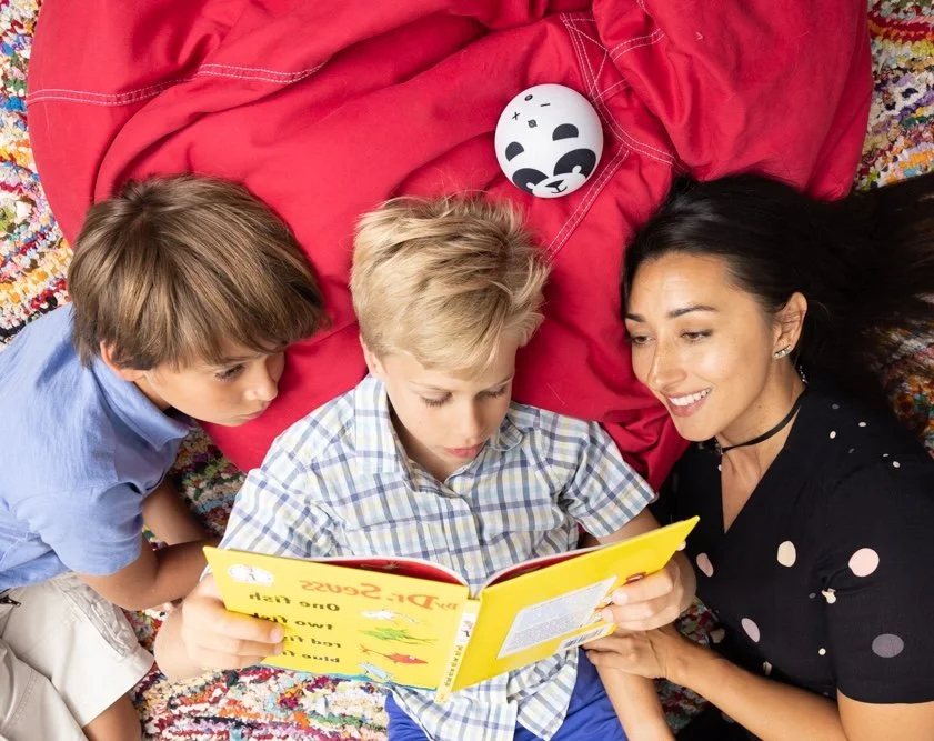 A woman and two young boys lying on a colorful carpet, reading a yellow children's book. The boy in the middle has blonde hair, the boy on the left has light brown hair, and the woman has dark hair. There is an Amazon Alexa  device nearby.