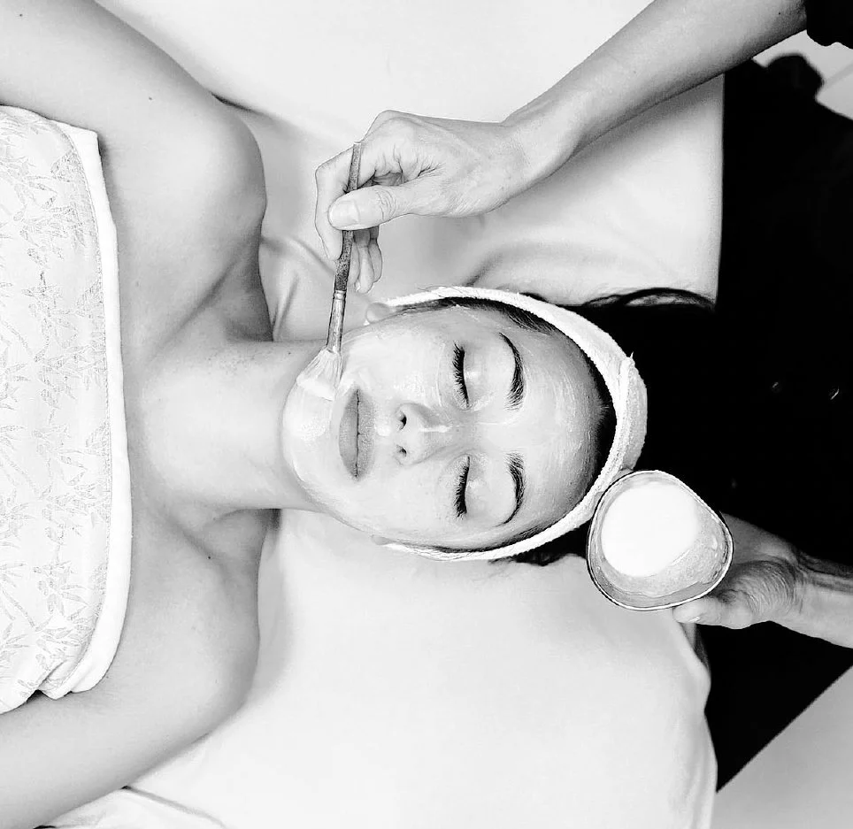 A woman lying on her back with her eyes closed, receiving a facial treatment. A beautician applies a cream with a brush and holds a small container of cream.