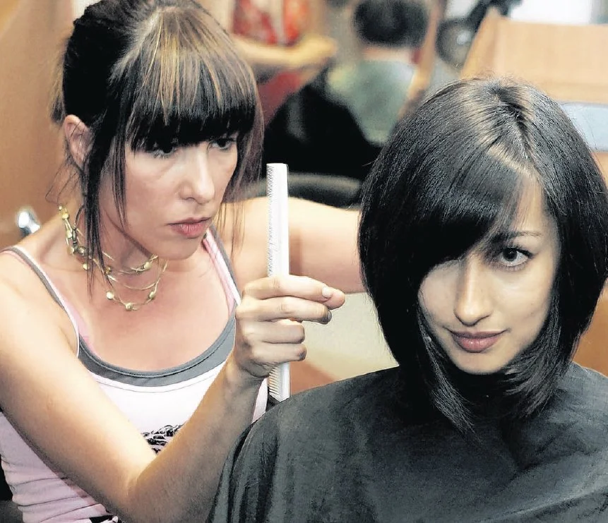 A woman with dark, straight, shoulder-length hair getting a haircut at a salon, while another woman with dark hair and bangs works on her hair.