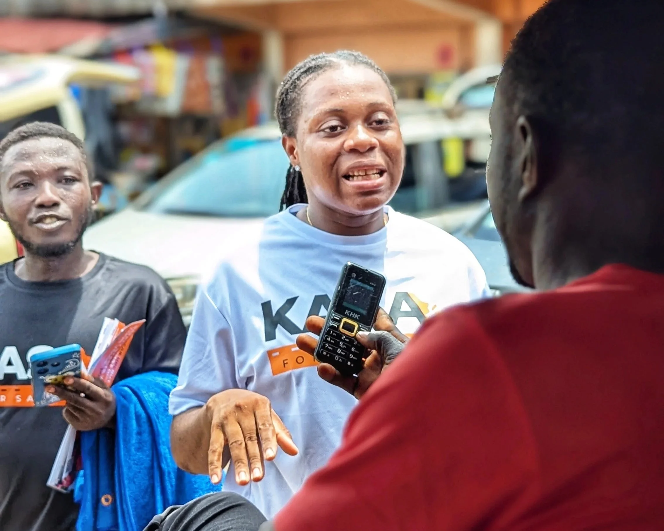 A woman showing her hand to a man holding a phone while two young men look on. The woman wears a white shirt with an orange badge, and her hand appears injured or injured with a bandage or scrape. The background shows parked cars and a wooden structure.