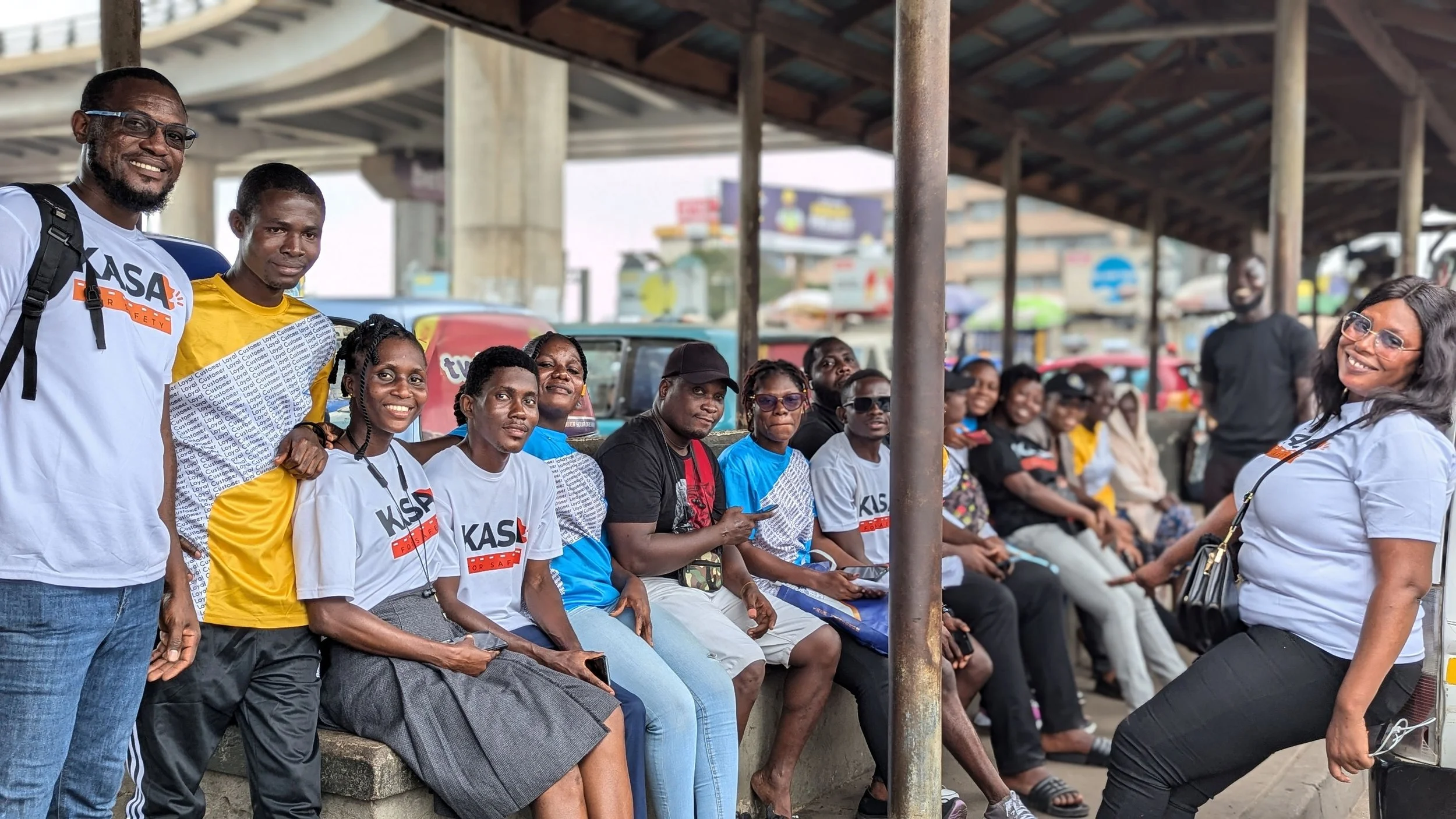A group of people sitting and standing on a bus stop or platform. Some of them are wearing white t-shirts with the letters KASA on them. The background shows cars and a cityscape. Two women and three men are standing, while others are seated and waiting.