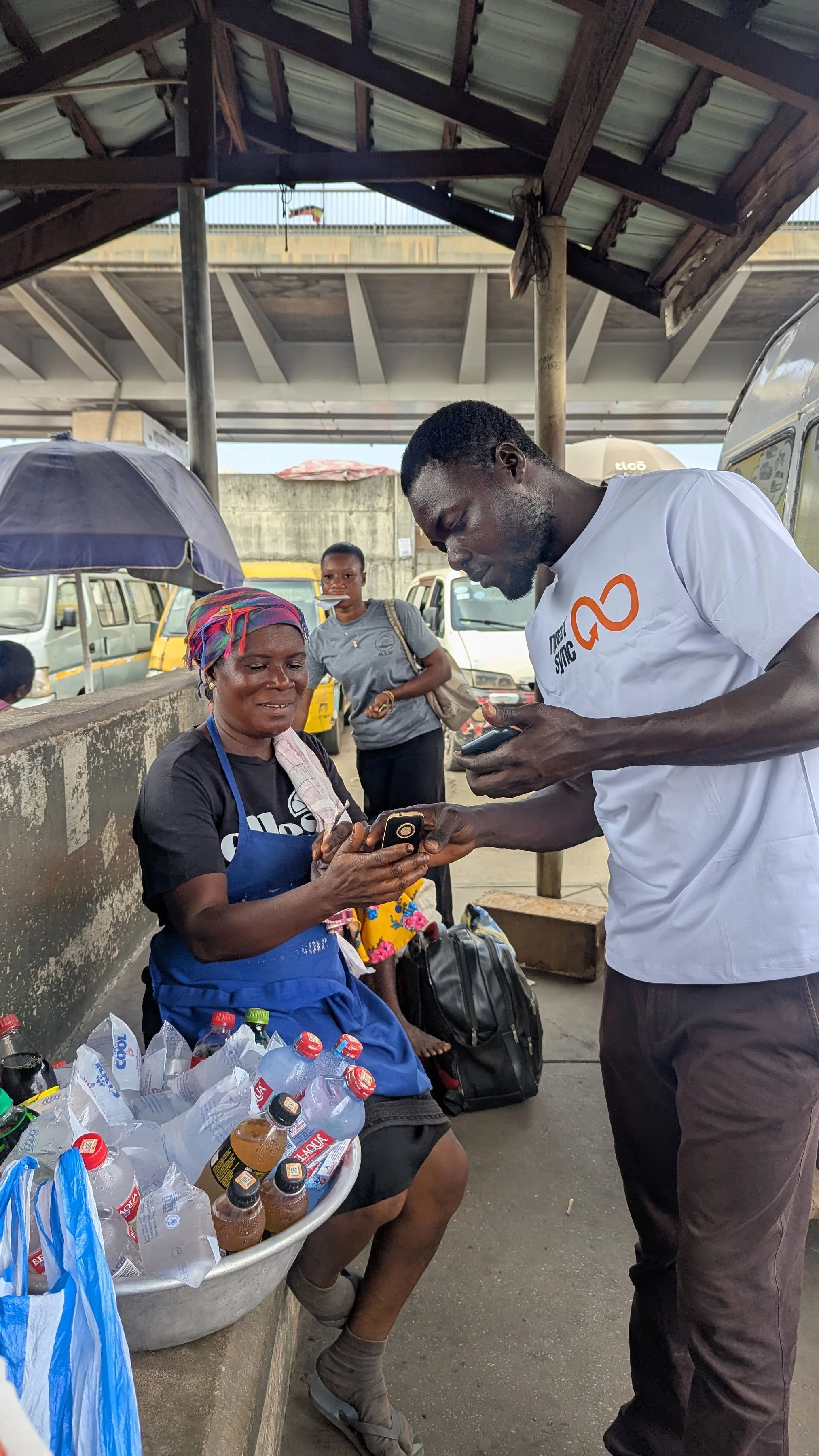 A woman sitting by a street vendor stall with bottles of water and drinks, showing her a phone while a man stands beside her. Another woman in the background holds a bag and looks on.