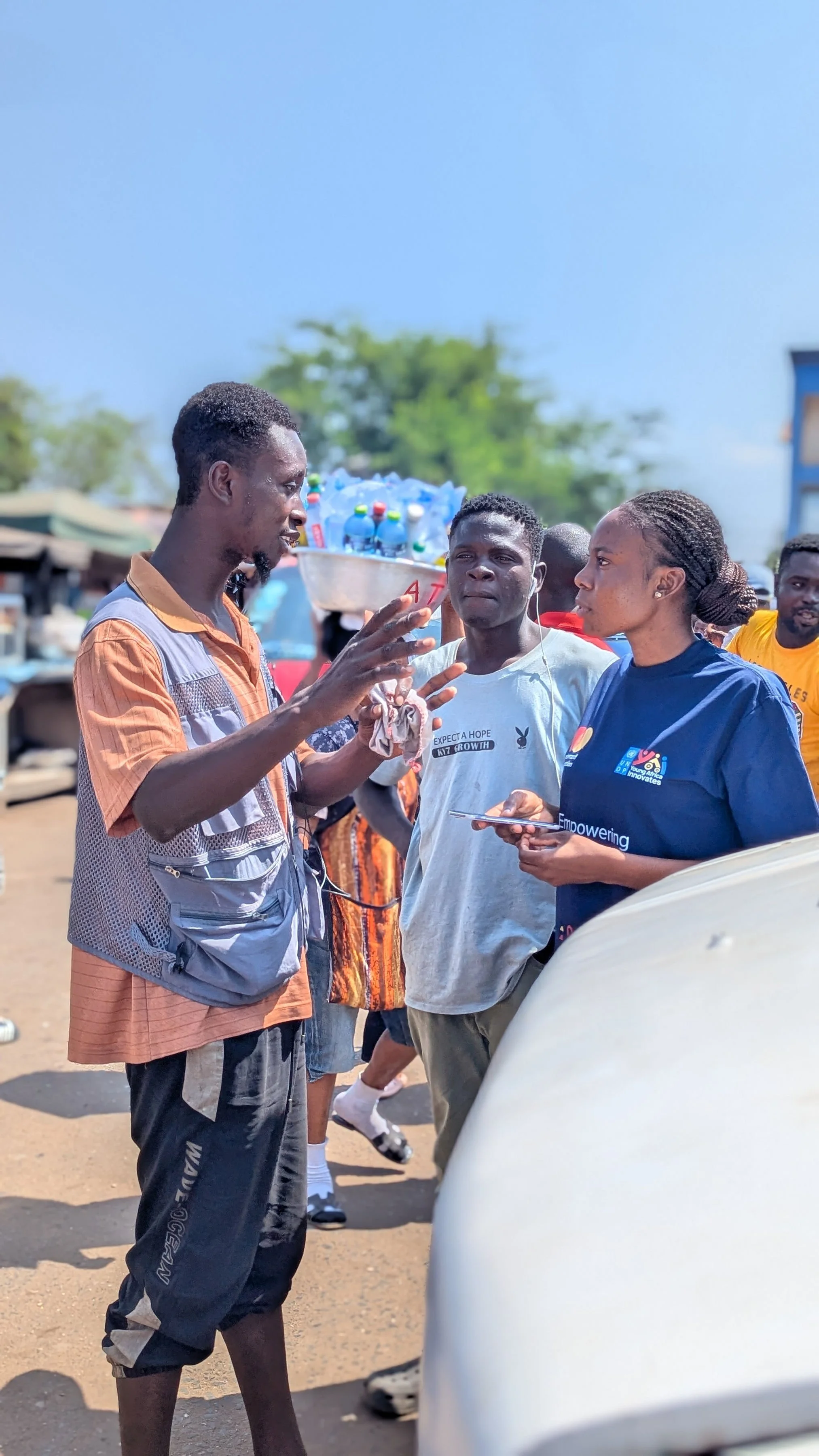 A group of people engaged in conversation outdoors, with one man gesturing with his hands while talking to a woman holding a phone. Others look on, some listening attentively. The background shows trees, market stalls, and a clear blue sky.
