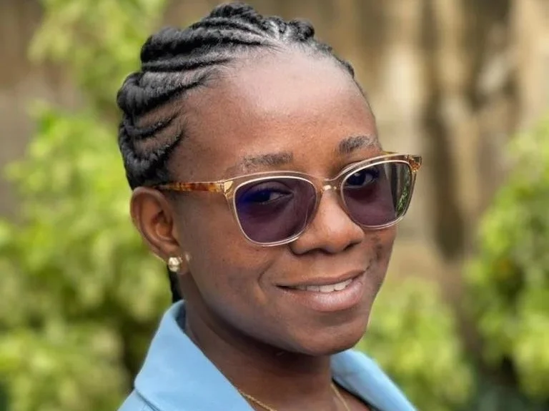 Close-up of a woman wearing sunglasses, an elegant light blue shirt, earrings, and a necklace, with a blurred green outdoor background.