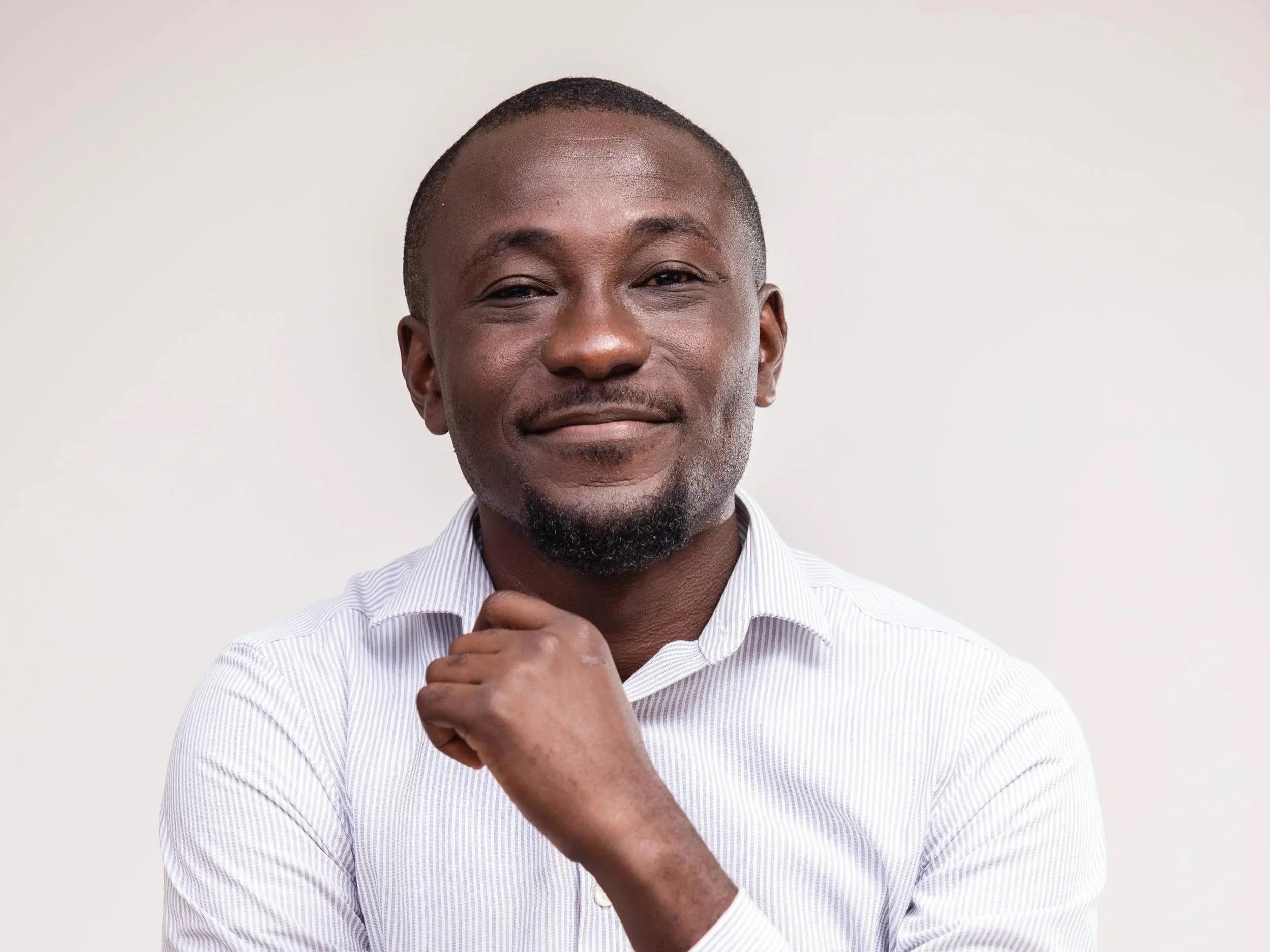 Close-up of a smiling African American man with short hair, wearing a light striped shirt, standing against a plain light background.