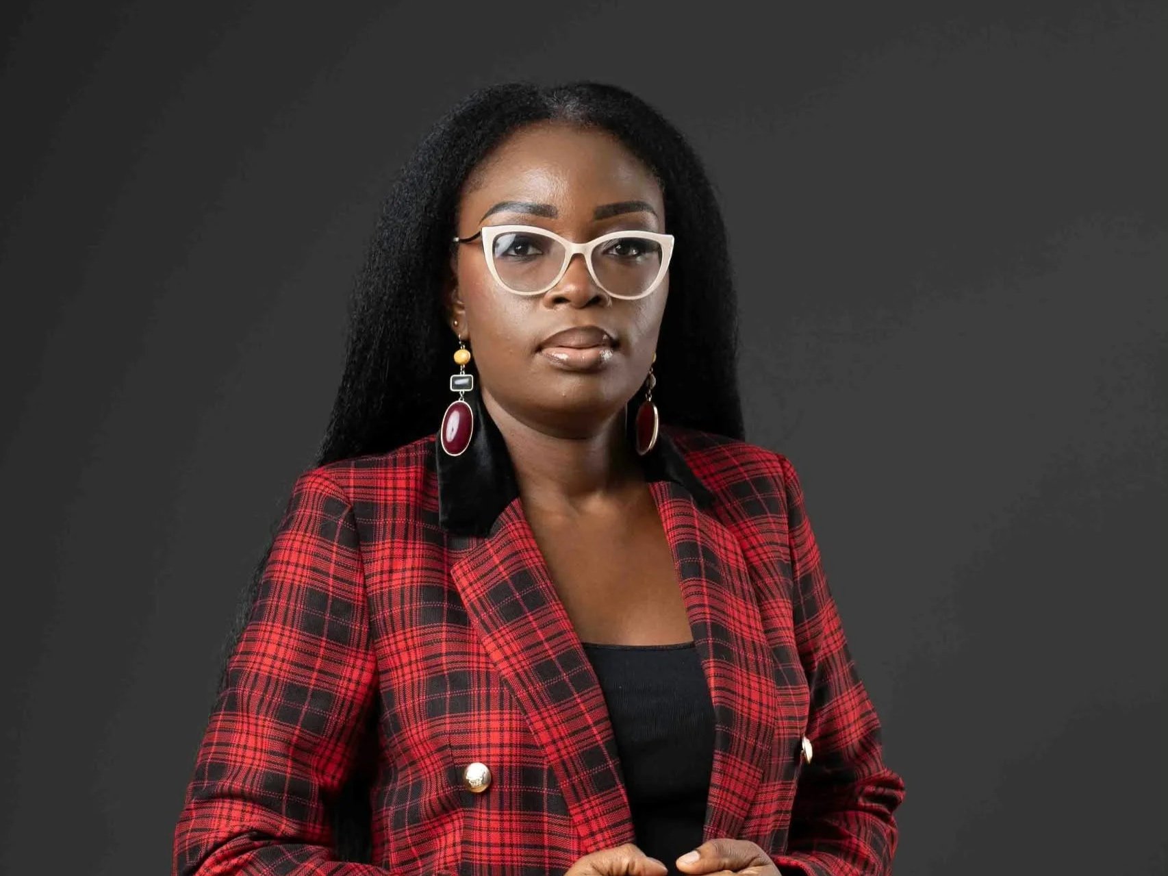 A woman wearing white-rimmed glasses with long black hair, dressed in a red plaid blazer with black accents and large earrings, standing against a dark gray background.