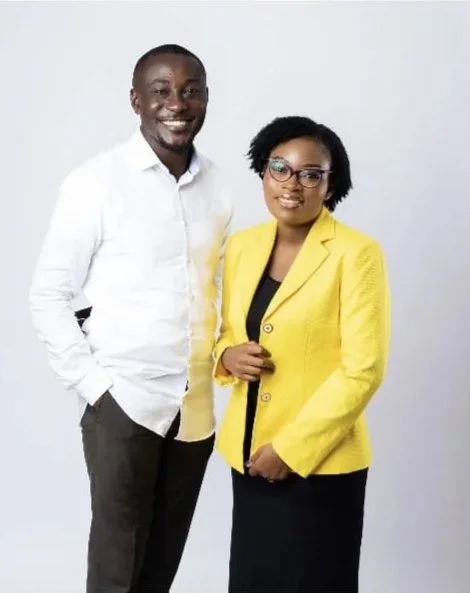 A man and woman standing together against a plain white background. The man is wearing a white shirt and gray pants, smiling with his hands in his pockets. The woman is wearing a yellow blazer over a black top and skirt, with glasses, and is smiling with one hand holding her blazer lapel.