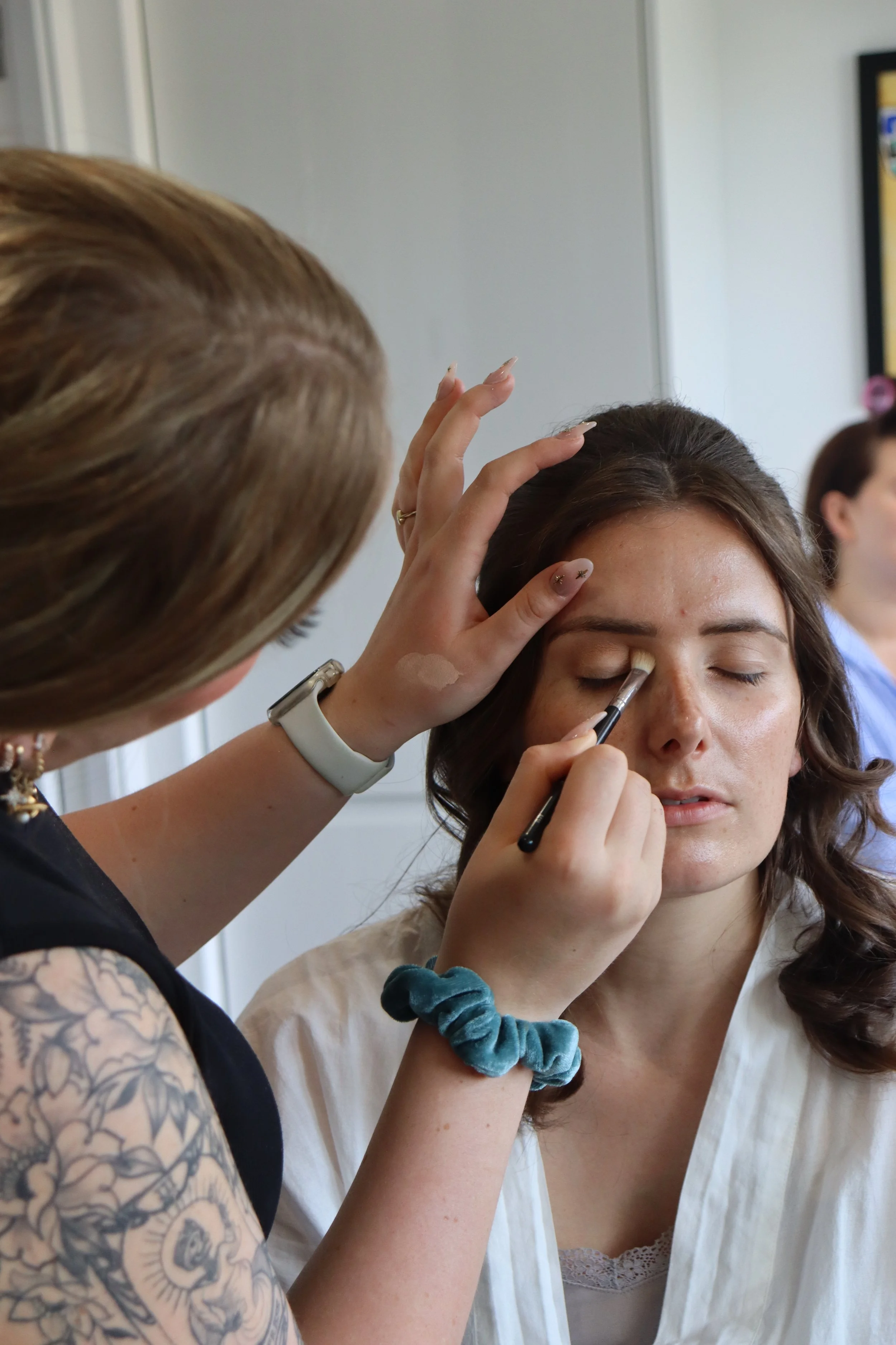 A person with dark brown hair is having makeup applied to their eyelid by another person with short, light brown hair and a tattoo sleeve on their left arm. The makeup artist is holding a brush near the person's eye while using their other hand to steady their head.