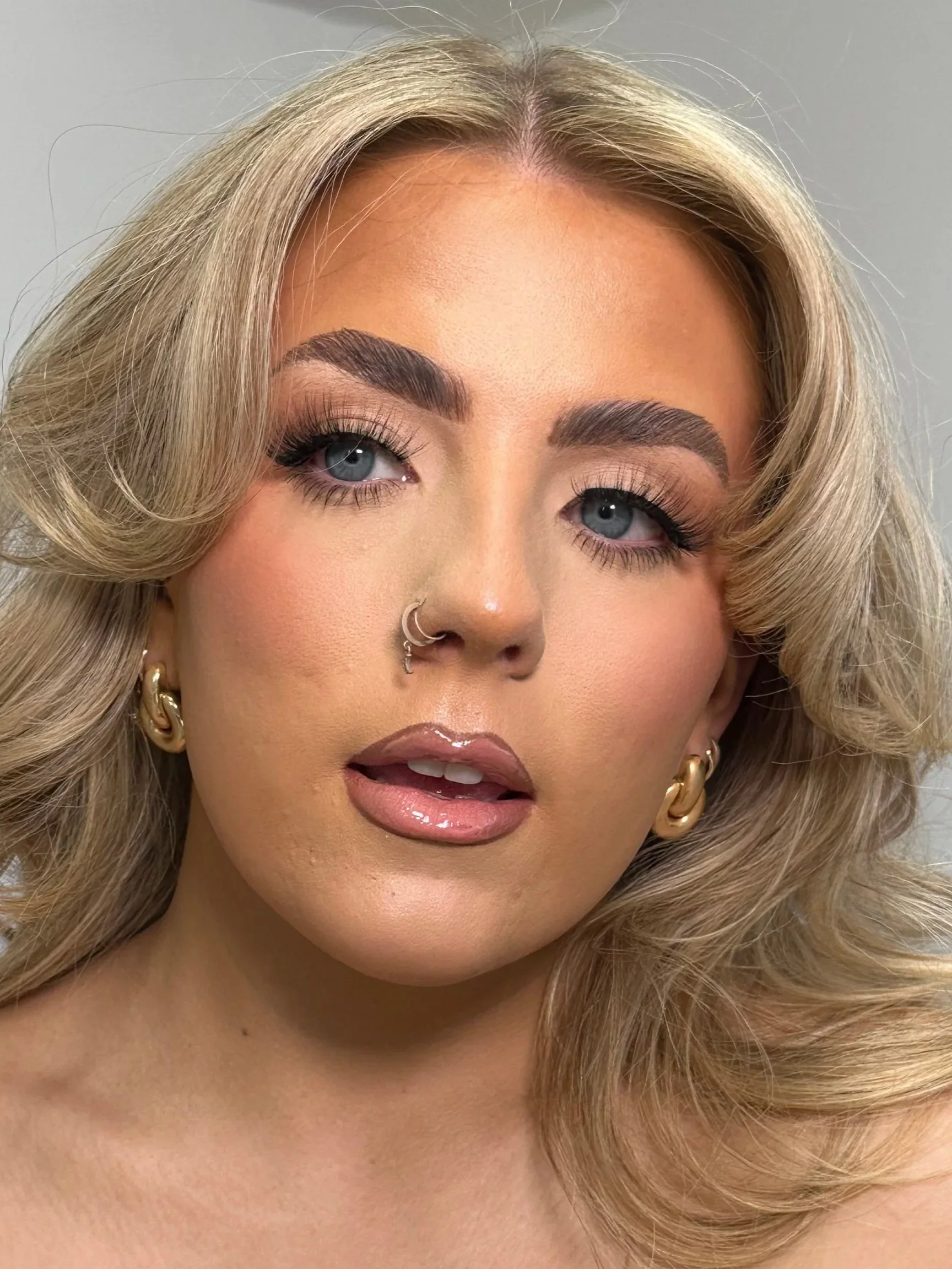 Close-up of a woman with blonde, wavy hair, blue eyes, makeup, and gold hoop earrings, looking to the side.