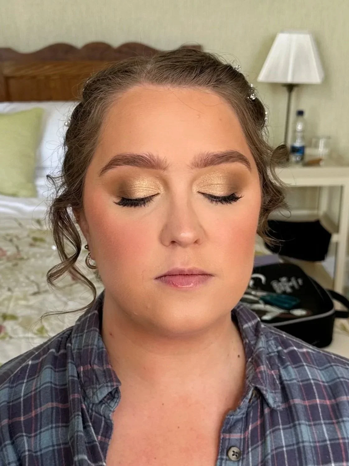 A woman with closed eyes showing gold eyeshadow and makeup, wearing a plaid shirt, indoors with a bed, lamp, and a water bottle in the background.