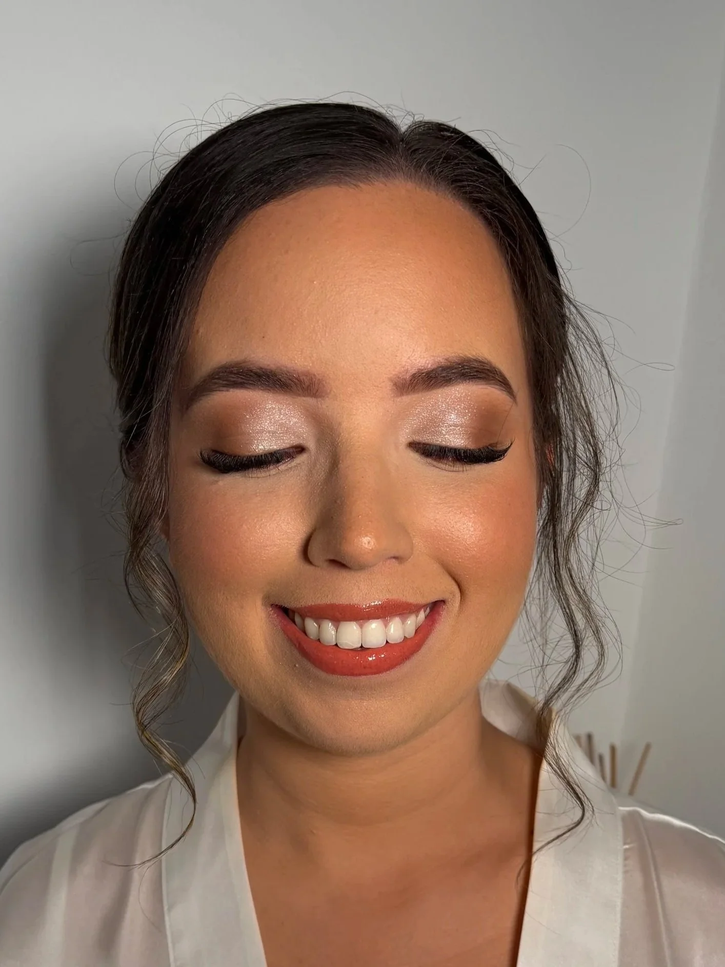 A woman with dark brown hair styled in loose curls, closed eyes with shimmery eyeshadow, and a broad smile showing her teeth. She is wearing a white top and appears to be happy and relaxed.