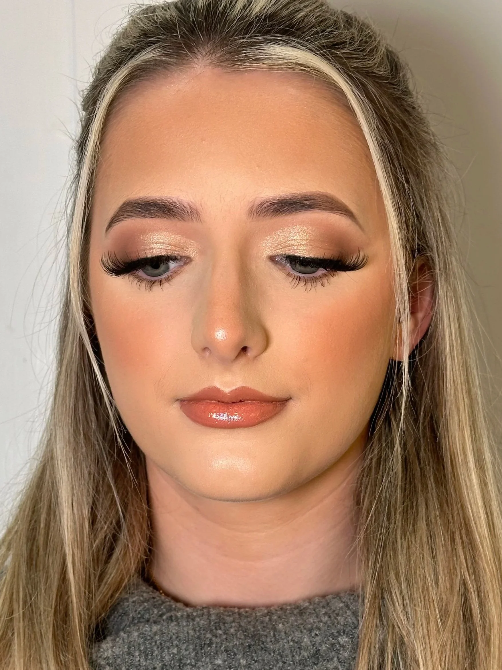 A close-up of a woman's face with makeup, including eyelash extensions, eyeshadow, and glossy lips. She has blonde hair and is wearing a gray top.