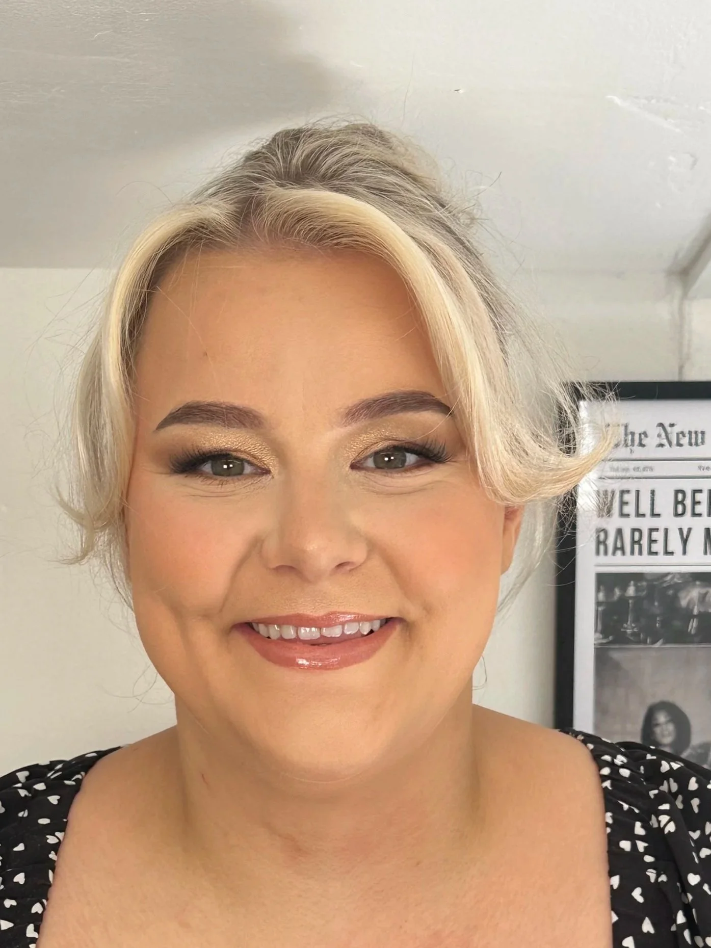 Close-up of a smiling woman with blonde hair styled in soft waves, wearing makeup with gold eyeshadow and nude lipstick, dressed in a black top with white hearts, standing indoors with a framed newspaper or poster on the wall behind her.