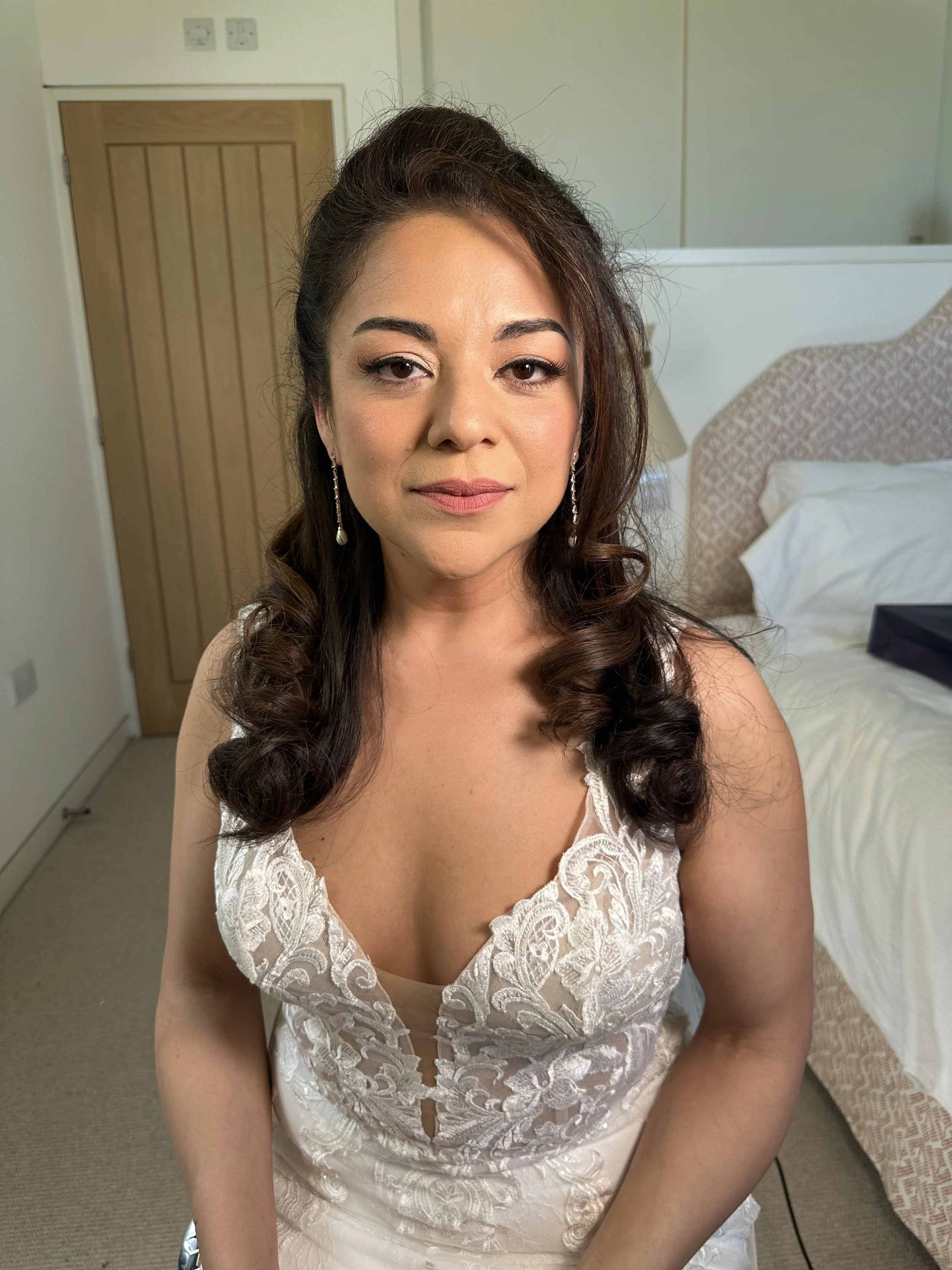 Woman in a white lace dress with pearl earrings, sitting in a room with a bed and wooden door, looking at the camera.