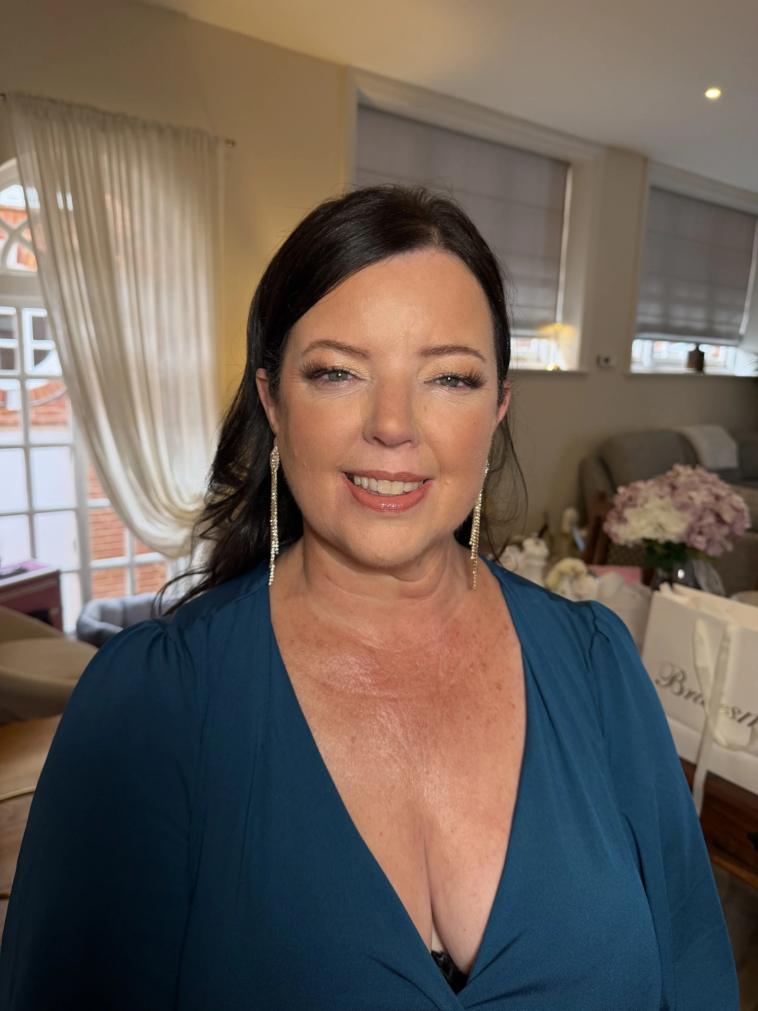 A woman with dark hair and earrings wearing a blue dress, smiling in a cozy room with white curtains, windows, and floral decorations in the background.