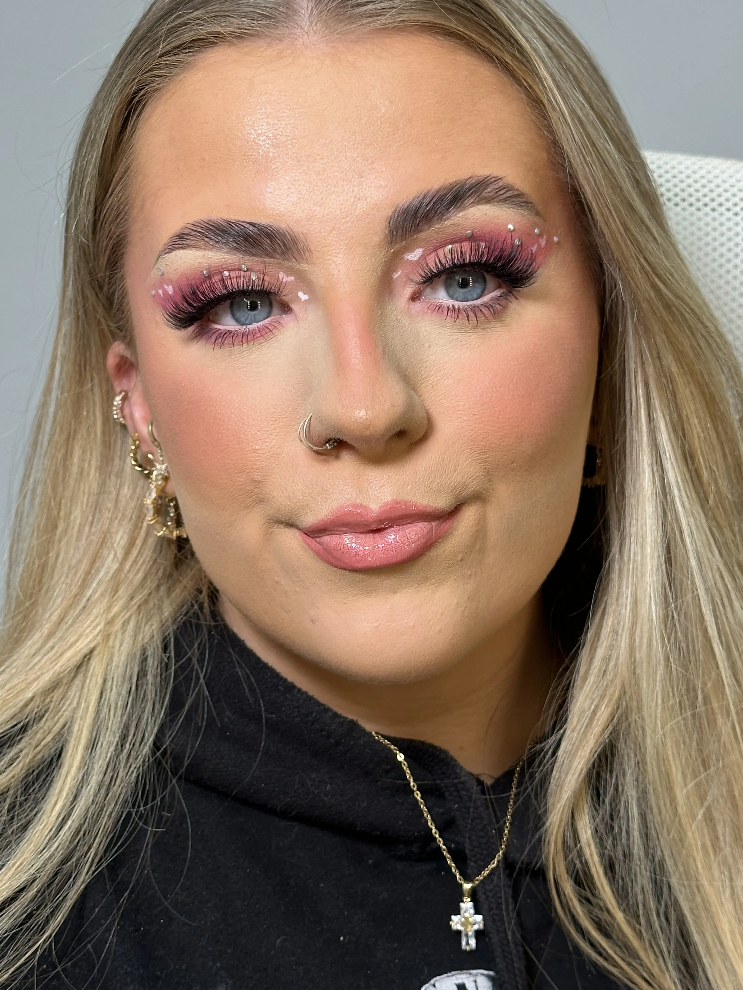 Close-up of a woman with blonde hair and blue eyes, wearing pink eyeshadow with white dots, false lashes, and a septum piercing, dressed in a black hoodie with gold jewelry.