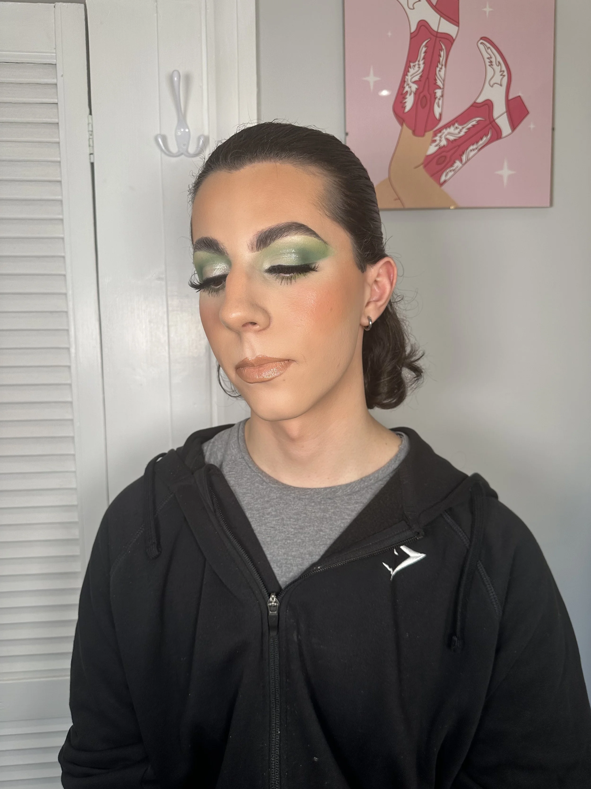 A woman with closed eyes, wearing bold green eyeshadow, shimmering lip gloss, and a black zip-up hoodie, standing in front of a white wall with a pink artwork of someone jumping or dancing wearing pink boots with white patterns.