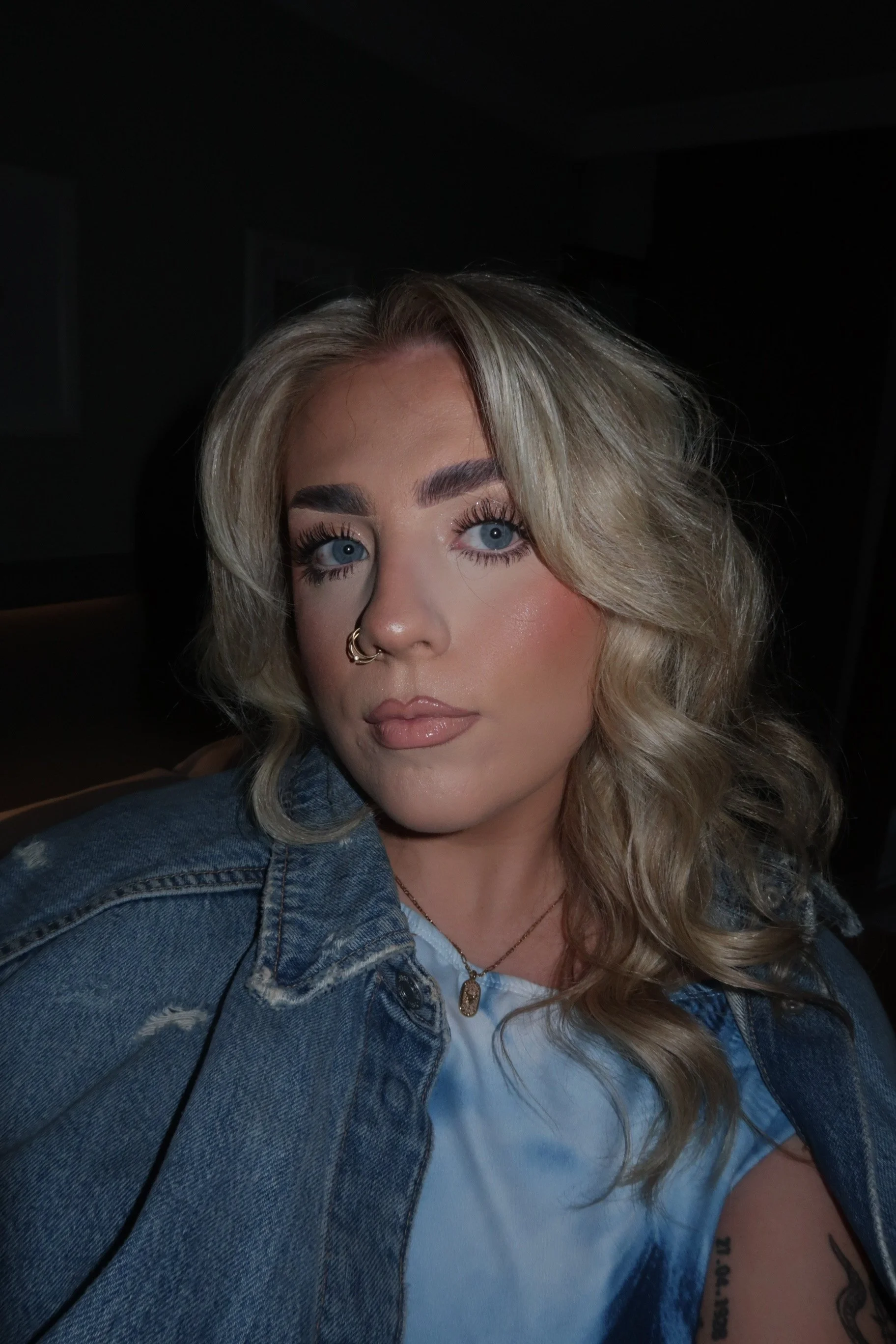 Close-up of a woman with wavy blonde hair, blue eyes, wearing makeup, a necklace, a denim jacket, and a white shirt, with a tattoo and a piercing on her nose. She is in a dimly lit environment.