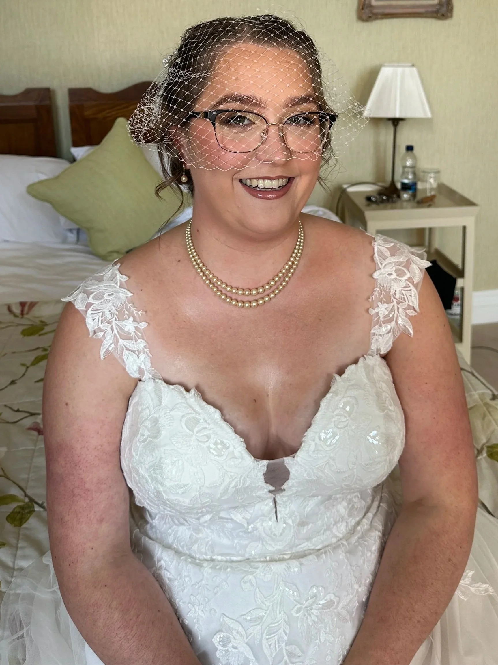 A smiling woman in a white wedding dress with lace details, wearing glasses, pearl necklace, and a birdcage veil, sitting in a room with a bed, pillows, and a nightstand with a lamp, water bottle, and other items.