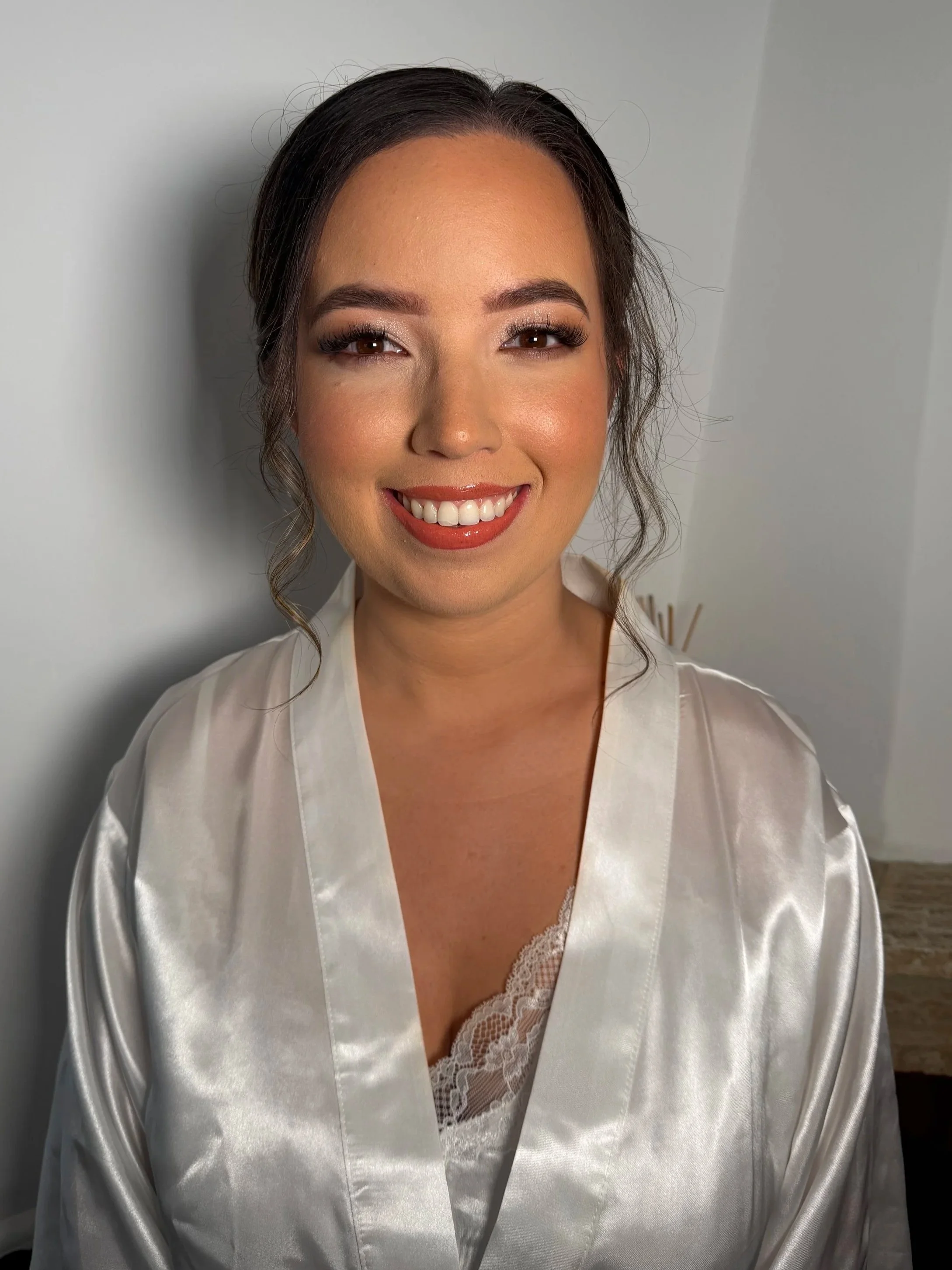 A woman with dark hair styled in loose waves, wearing a satin robe, and smiling in front of a plain wall.