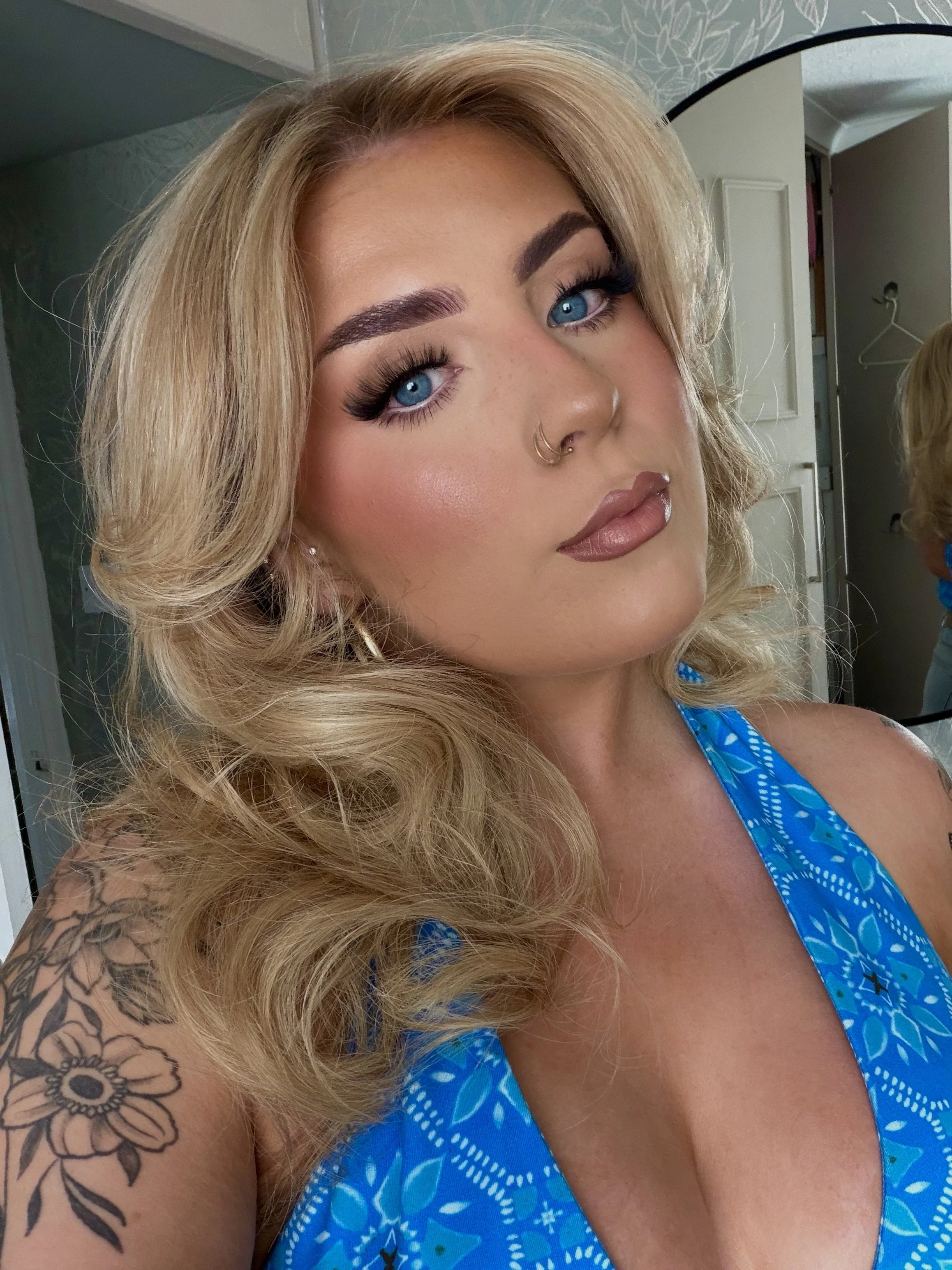 A woman with blonde, curly hair and striking blue eyes takes a close-up selfie. She has makeup, including dark mascara and brown lipstick, and is wearing a blue patterned top. A floral tattoo is visible on her upper arm.