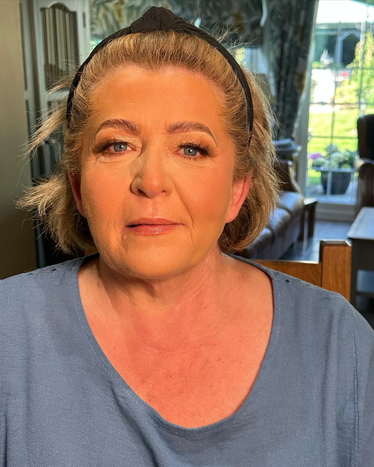 A middle-aged woman with short blond hair and blue eyes, wearing makeup including gold eyeshadow and lipstick, is sitting indoors near a window with a view of a green yard and furniture in the background.