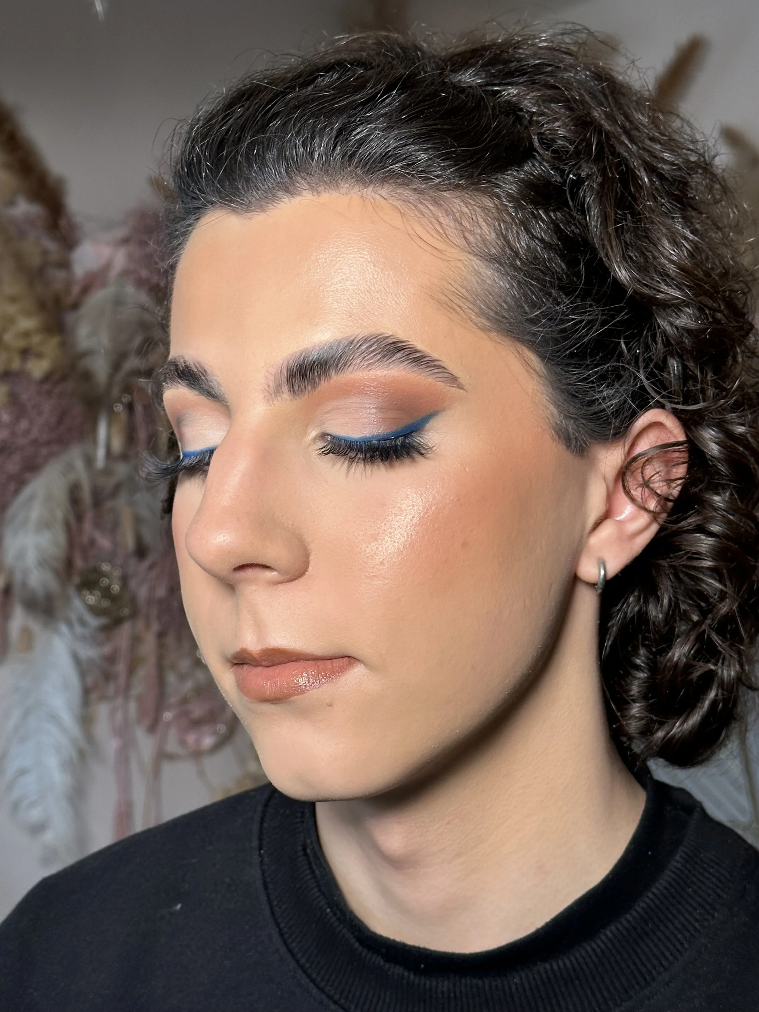 Close-up of a woman with short, curly black hair, closed eyes with blue eyeliner, and a neutral expression. She is wearing a black top and small hoop earrings.