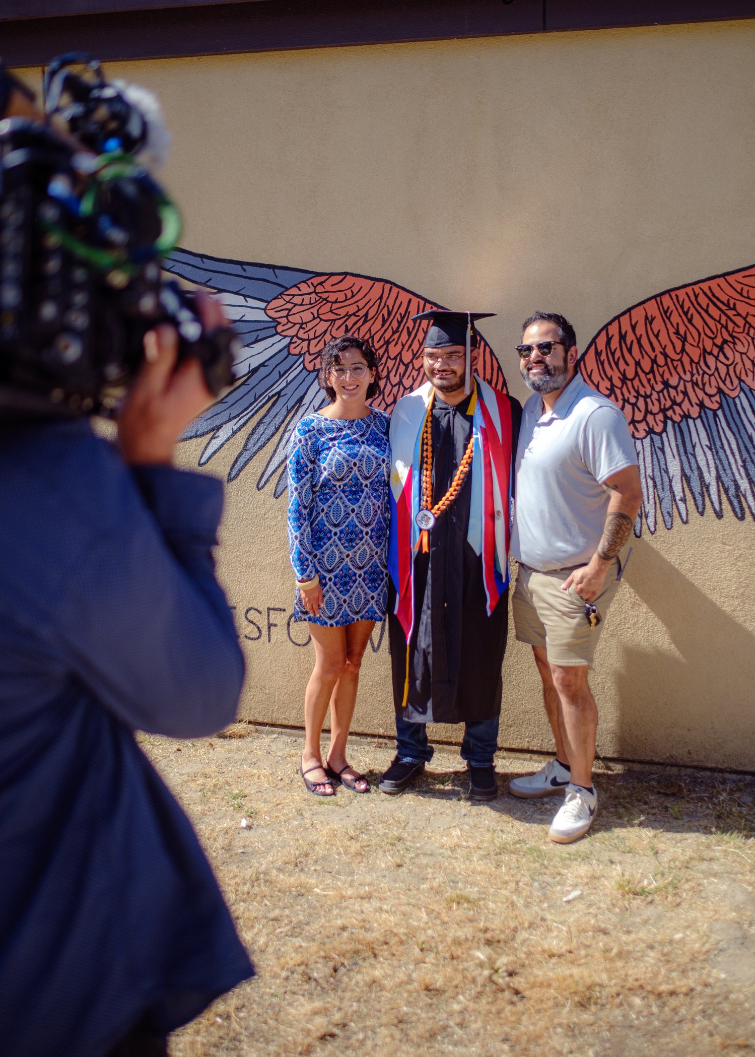 A man in a graduation cap and gown standing between a woman and a man in a white shirt, all smiling, with large wing murals on a beige wall behind them, camera in foreground capturing the moment.