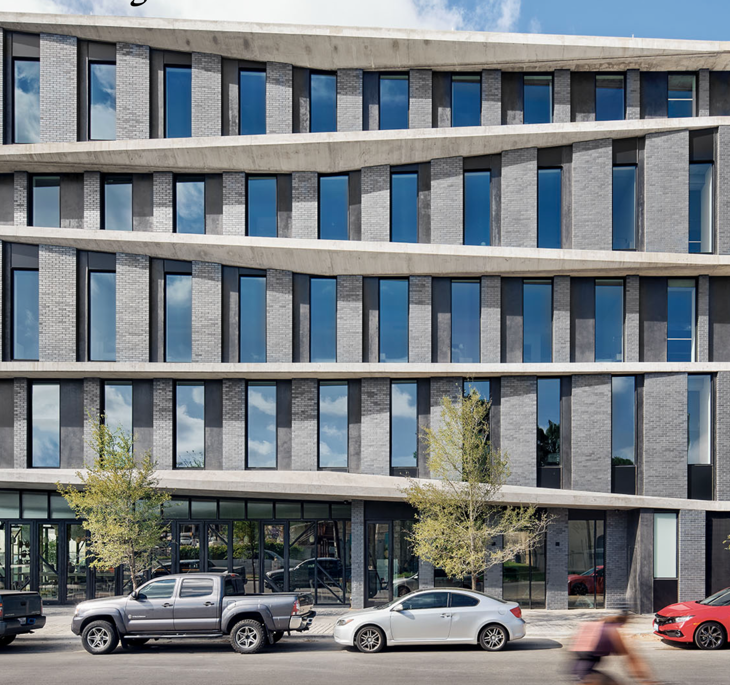 Modern multi-story building with large reflective windows and horizontal concrete accents, with trees and parked cars in front.