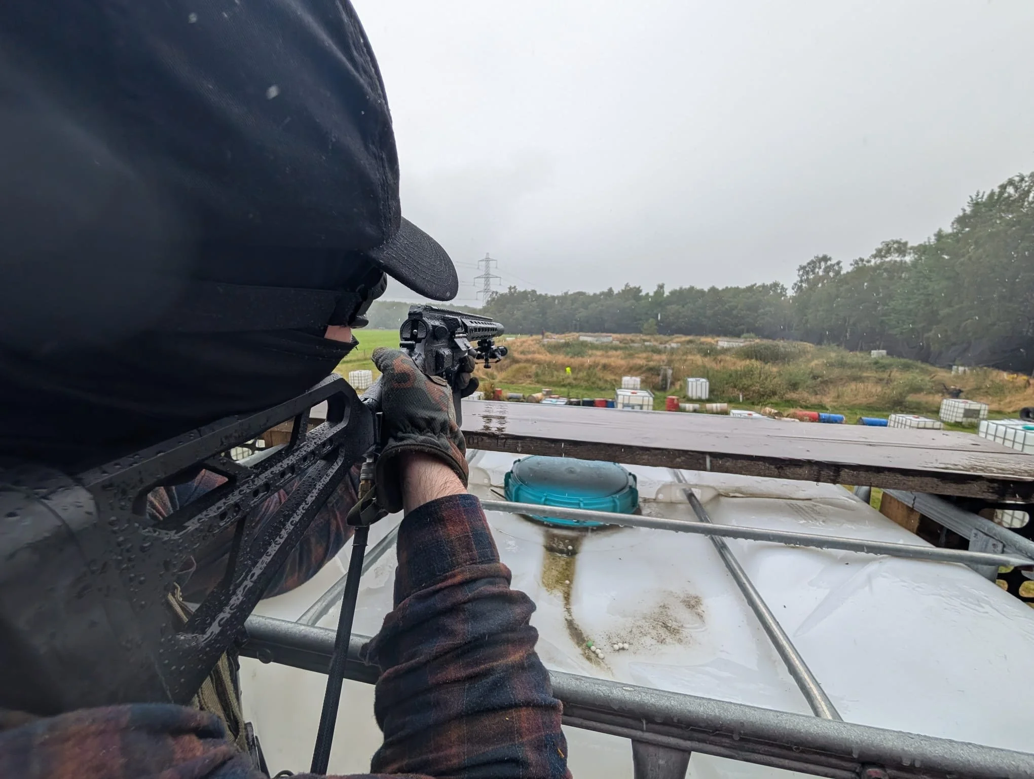 Person aiming a firearm from a shooting platform in an outdoor shooting range, with trees, barrels, and a cloudy sky in the background.