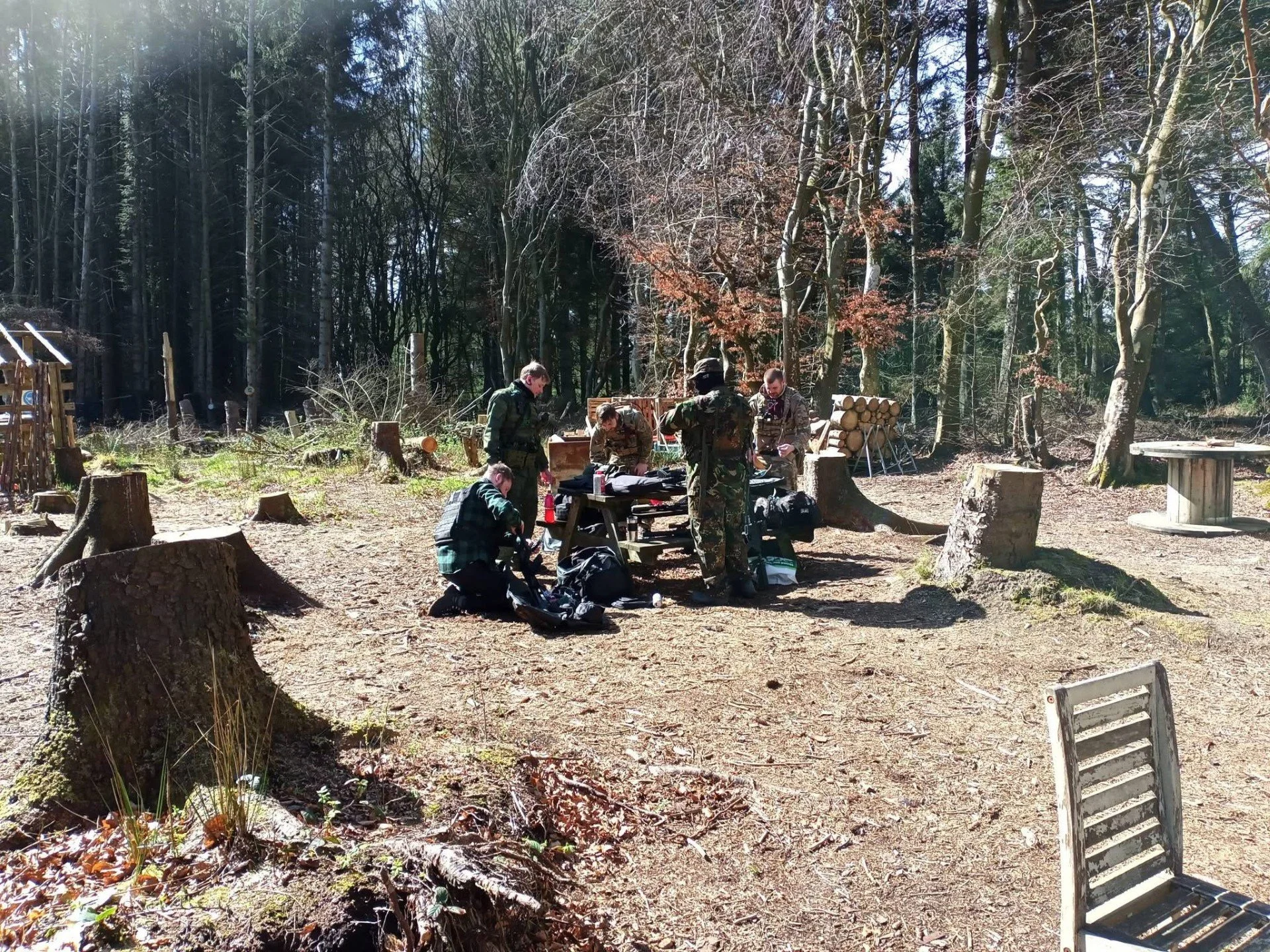 Group of soldiers in camouflage uniforms preparing at a wooden table in a forest clearing with cut tree stumps and logs, surrounded by tall trees.