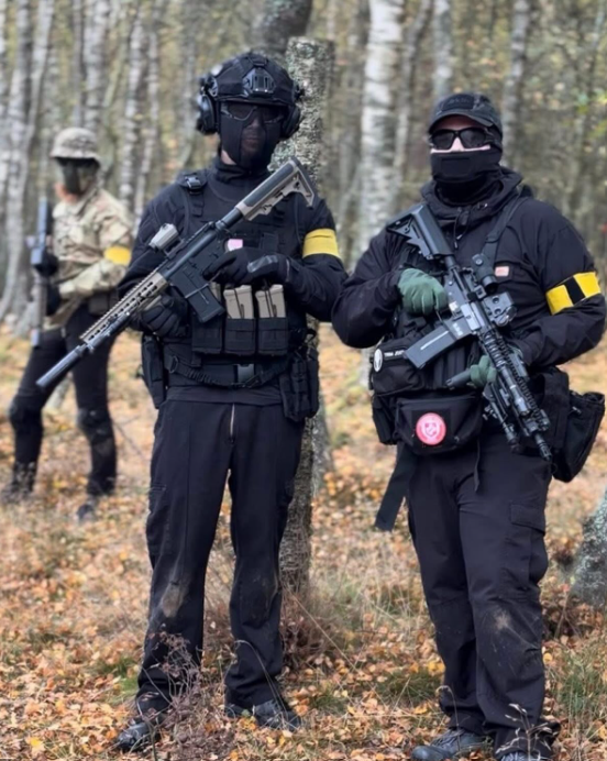 Three individuals dressed in tactical gear in a forest. The two in the foreground are facing the camera, wearing black outfits, masks, and holding rifles. The person in the background is wearing a beige outfit and mask, standing near a tree.